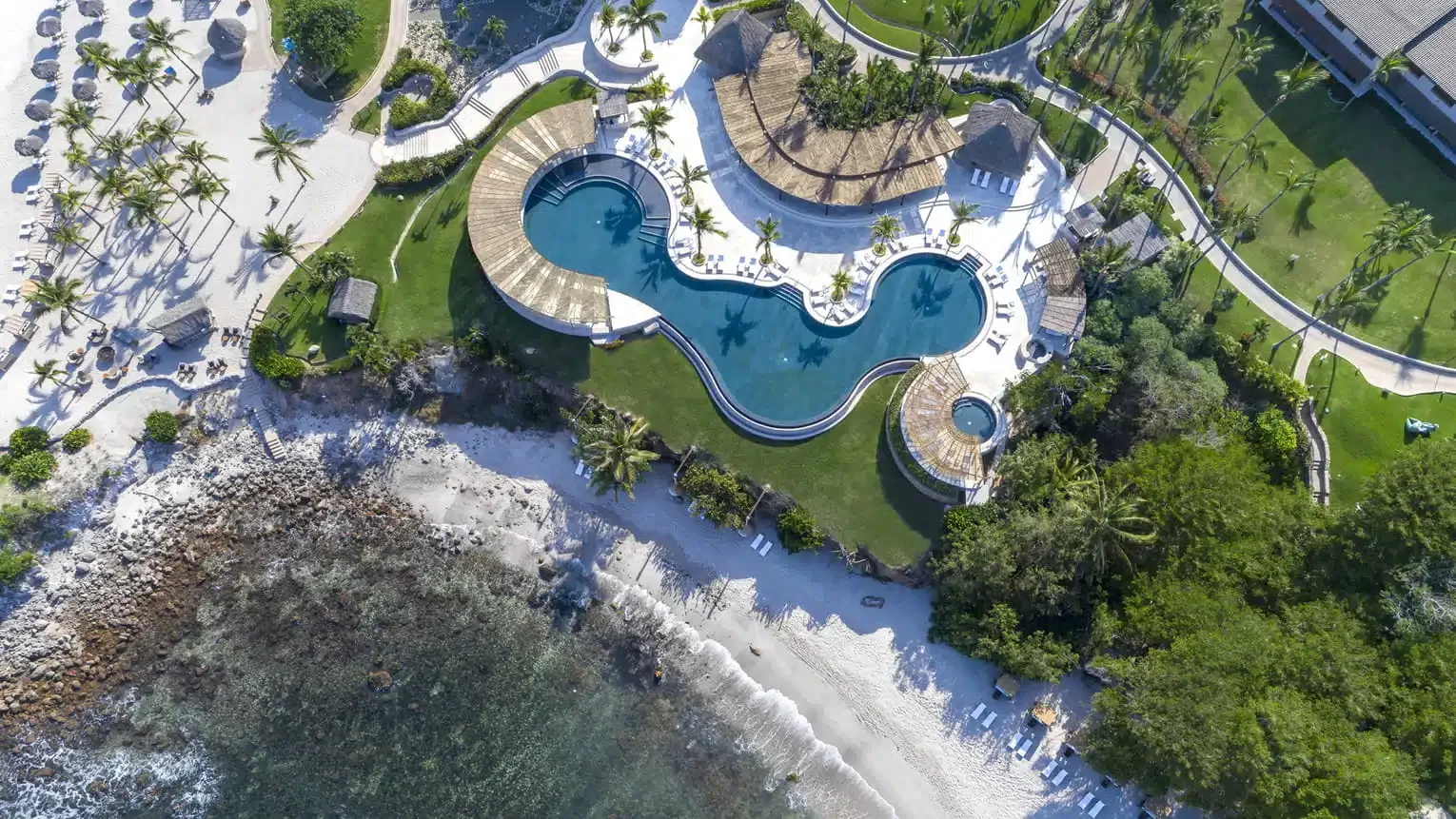 Aerial view of a resort pool area with curved pools, surrounding palm trees, lounge chairs, and a beach shoreline.