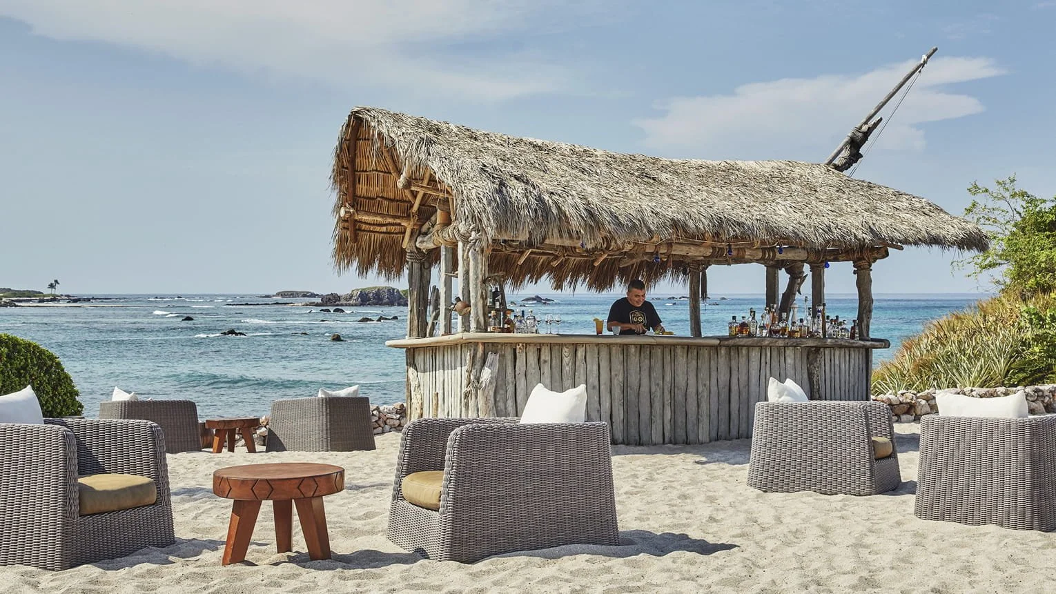 Beach scene with a tiki bar made of wood and thatched roof, surrounded by cushioned outdoor chairs, with the ocean and rocks in the background.