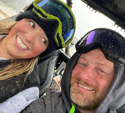 A smiling woman and man wearing helmets and gear, sitting in a side-by-side vehicle on a muddy outdoor trail.