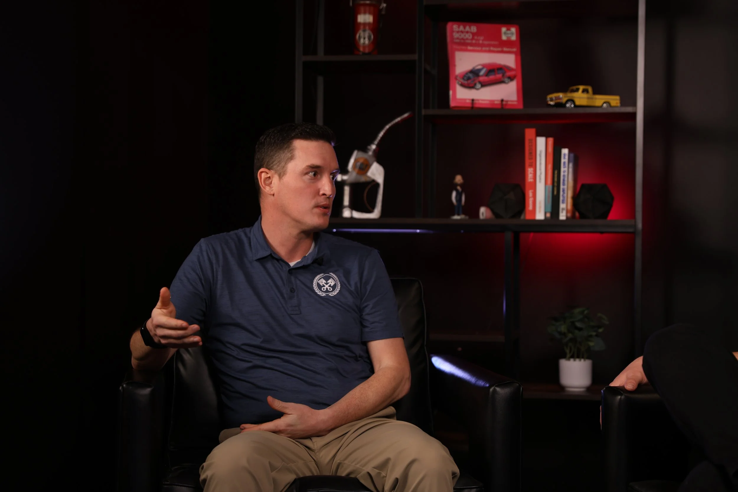 A man in a blue polo shirt seated in a black leather chair, gesturing with his right hand, inside a room with a black background and red-lit bookshelf behind him containing books and model cars.