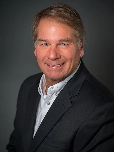 A professional headshot of a middle-aged man with gray hair, wearing a black blazer and white shirt, smiling in front of a dark background.