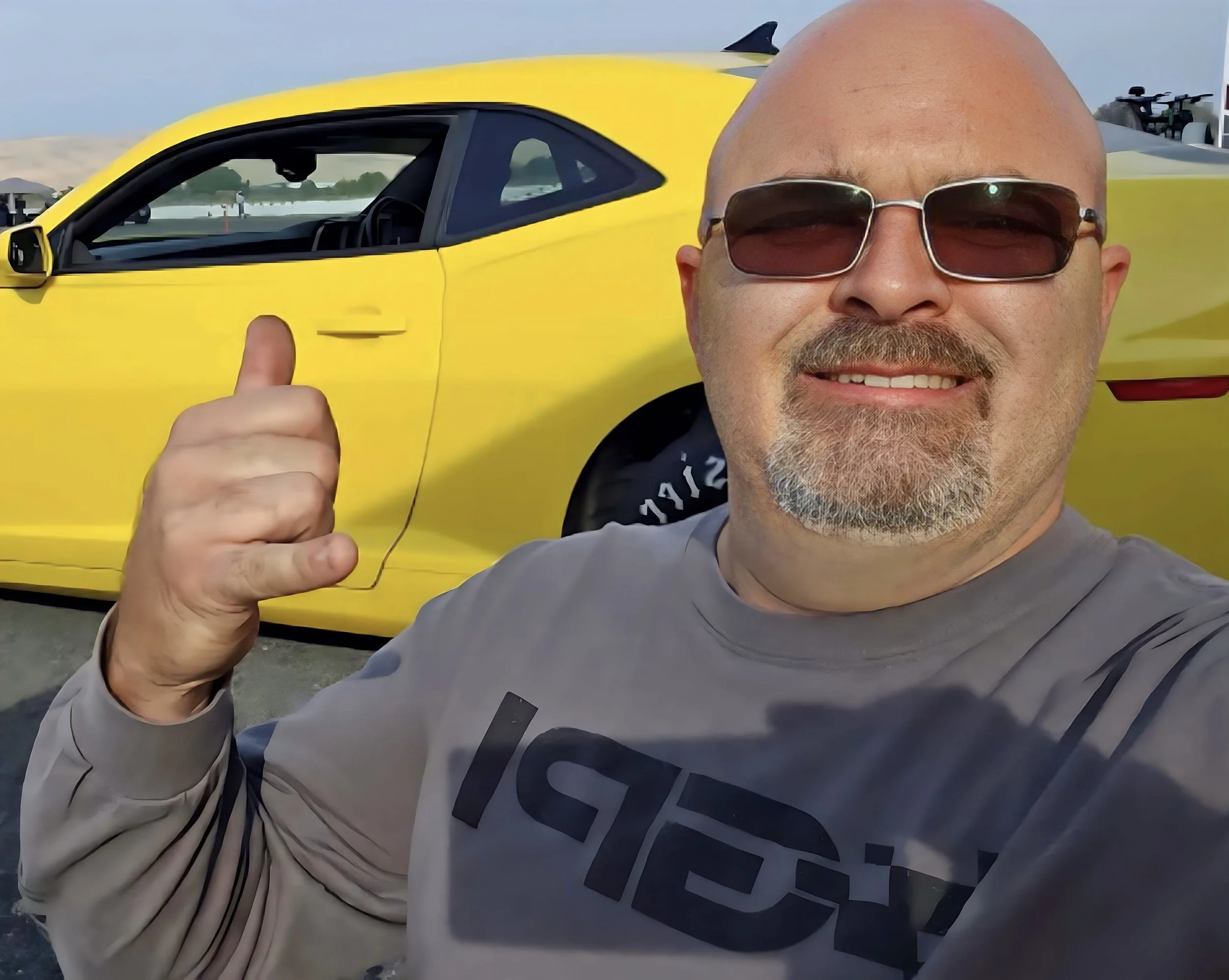 A smiling man with sunglasses and a goatee taking a selfie in front of a yellow sports car giving a thumbs up.