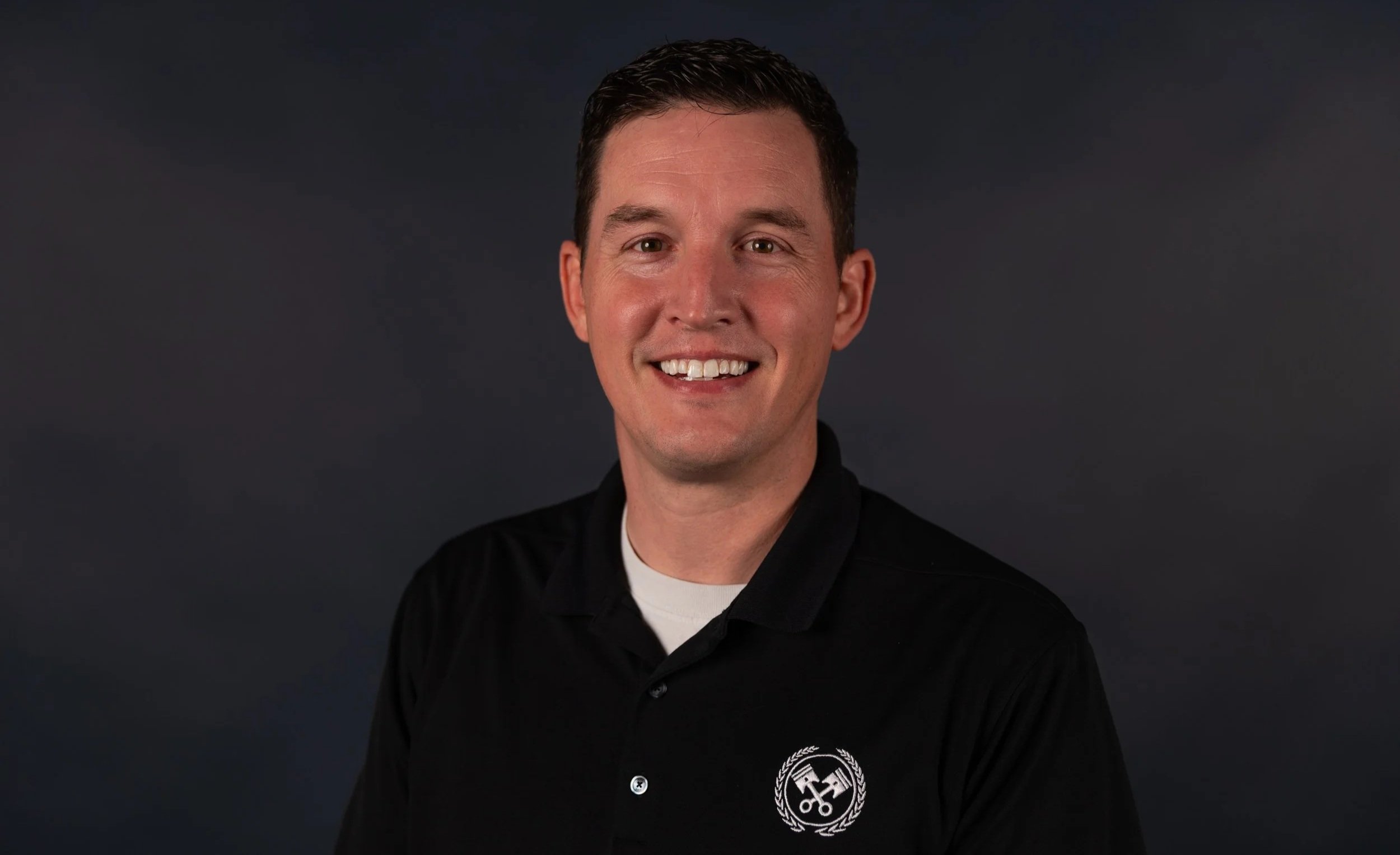 A smiling man with short dark hair wearing a light-colored checkered shirt.