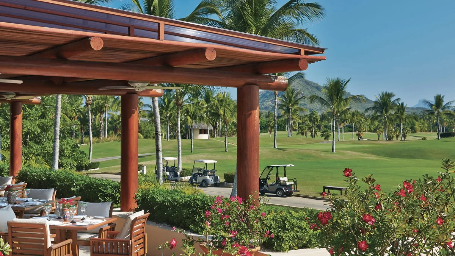 A sunny golf course viewed from a patio area with wooden chairs and a table in the foreground. Multiple golf carts are parked along a winding path, with lush green grass, tall palm trees, and distant mountains under a clear blue sky.