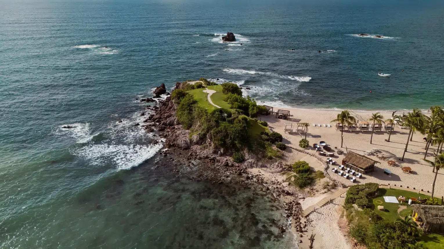 Aerial view of a tropical beach with a rocky outcrop in the ocean, green grassy area, palm trees, and beach cabanas.