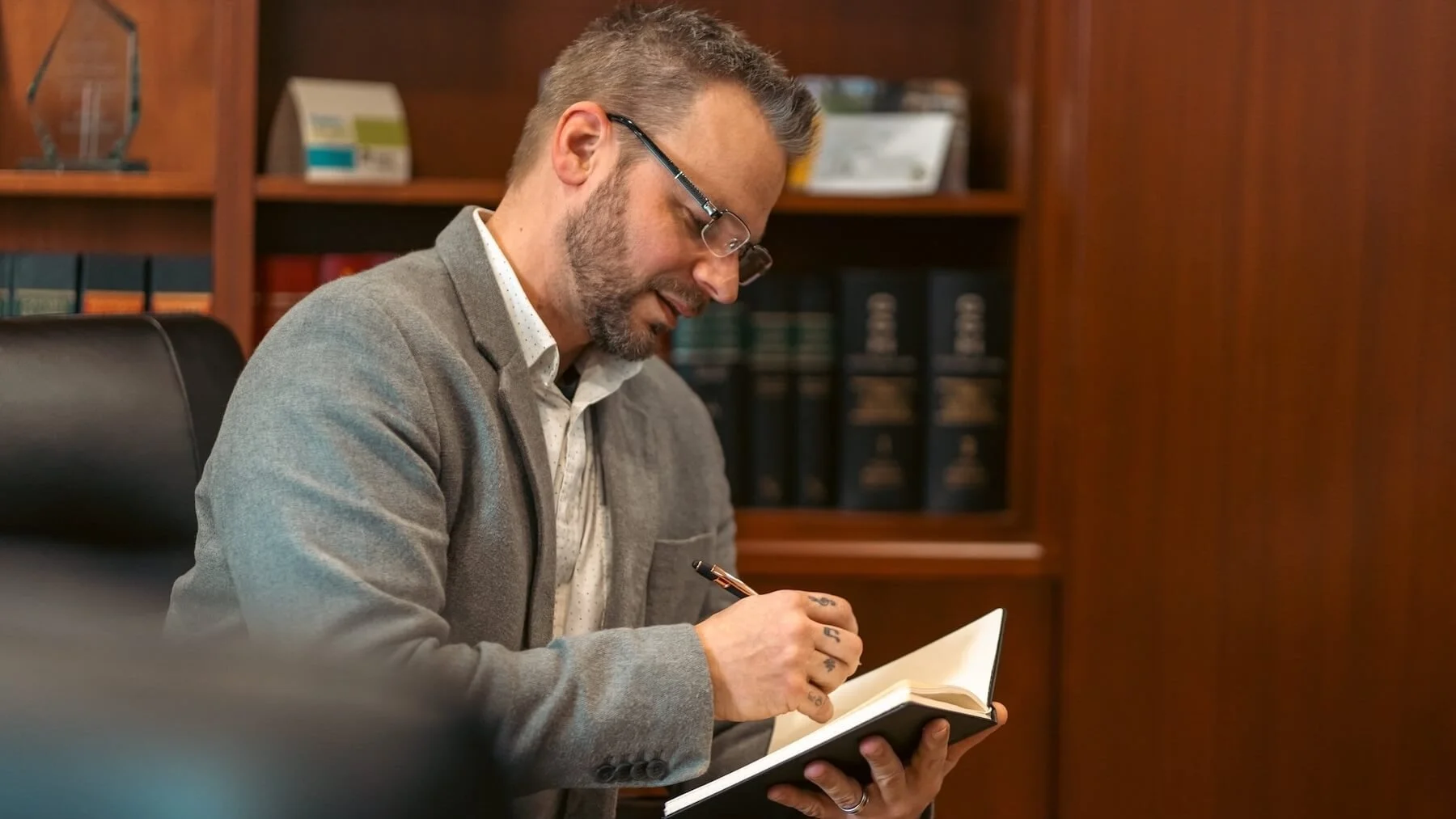 Man in glasses writing in a notebook in an office with wooden shelves and binders