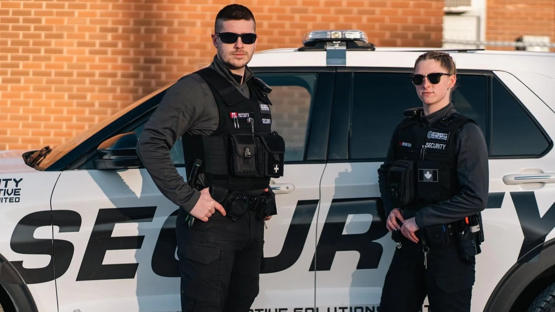 Two security personnel, a male and a female, standing in front of a police car with the word 'SECURITY' on it, wearing black uniforms and sunglasses.