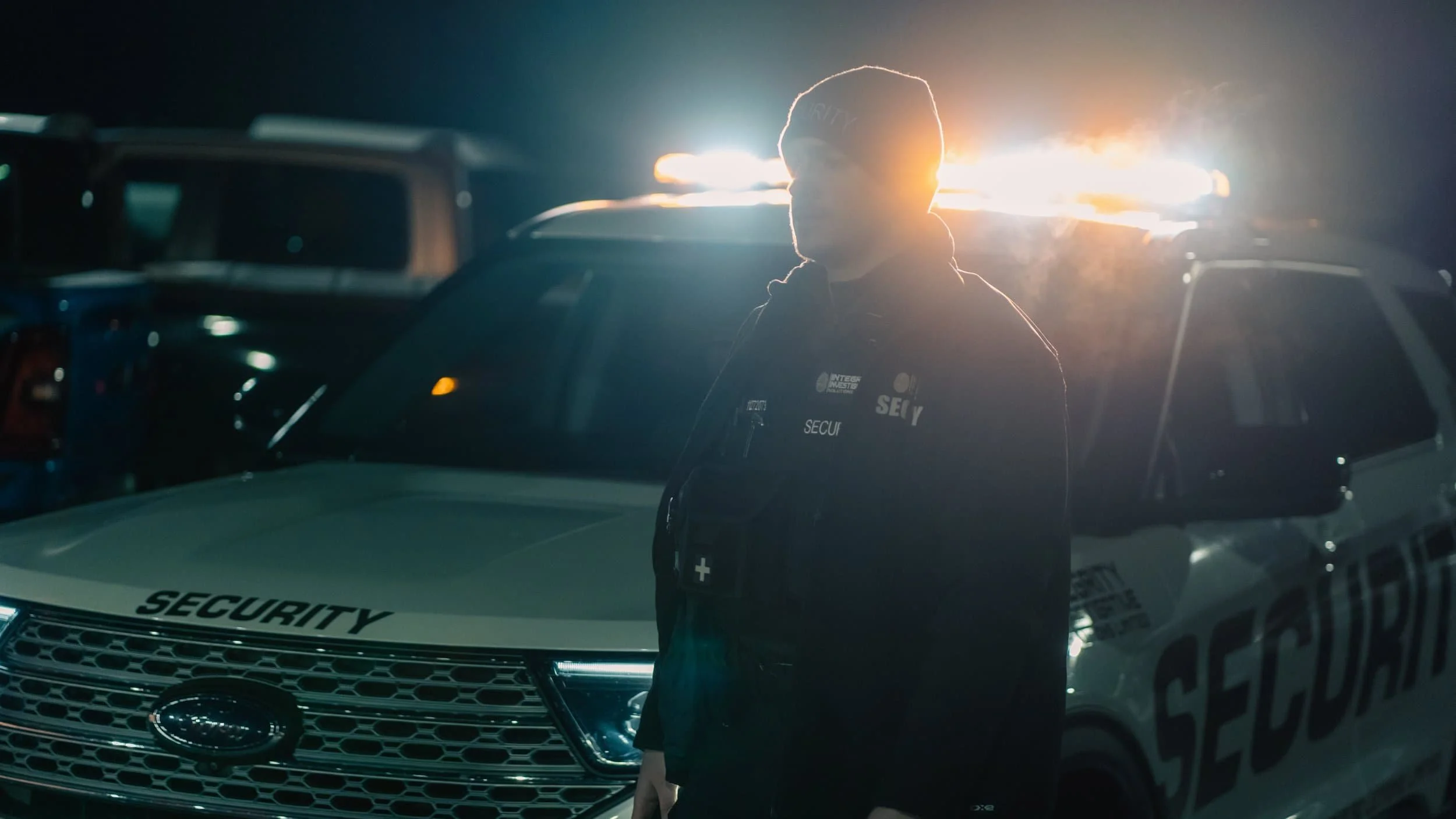 A security officer standing in front of a marked security vehicle with flashing lights on at night.
