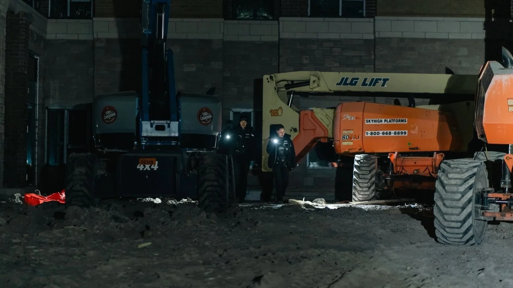 Nighttime construction scene with two large construction vehicles, a JLG lift and an orange telescopic boom lift, with two workers standing between them, illuminated by their flashlights.