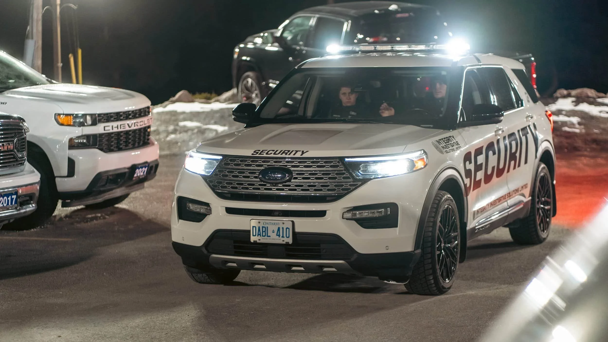 A white security police SUV with flashing lights parked on a nighttime street, with other parked cars around and snow on the ground.
