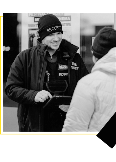 A smiling security guard in uniform holding a tablet and interacting with a person in a coat.