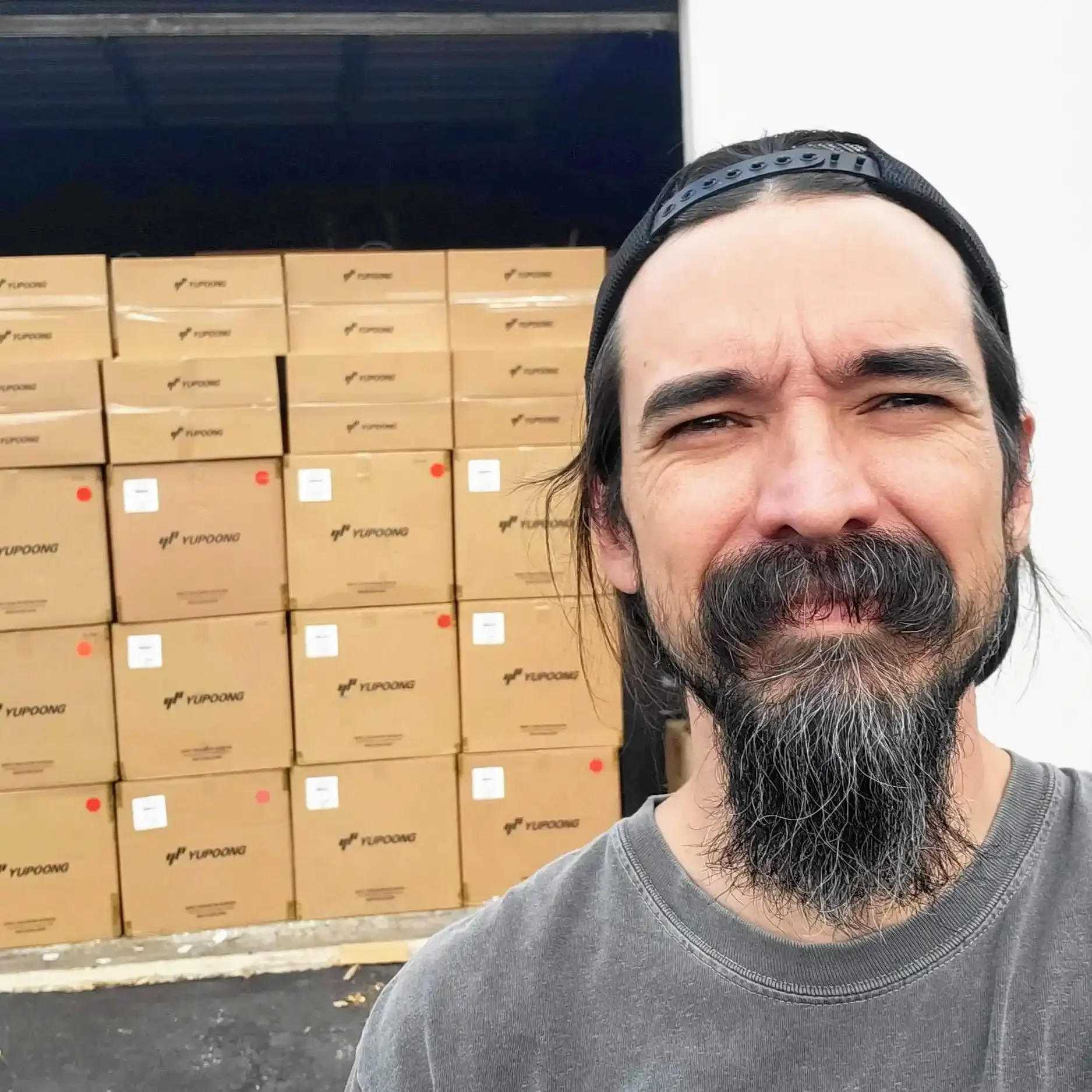 A man with a beard and mustache wearing a backward black cap and gray t-shirt taking a selfie in front of a stack of boxes in a warehouse.