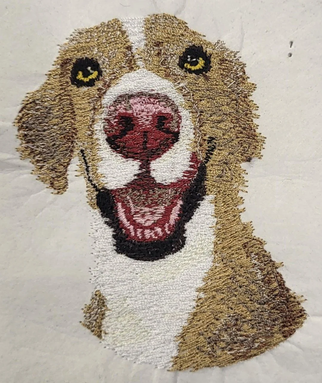 An embroidered image of a smiling dog with an open mouth, tan and white fur, and bright yellow eyes on a white background.