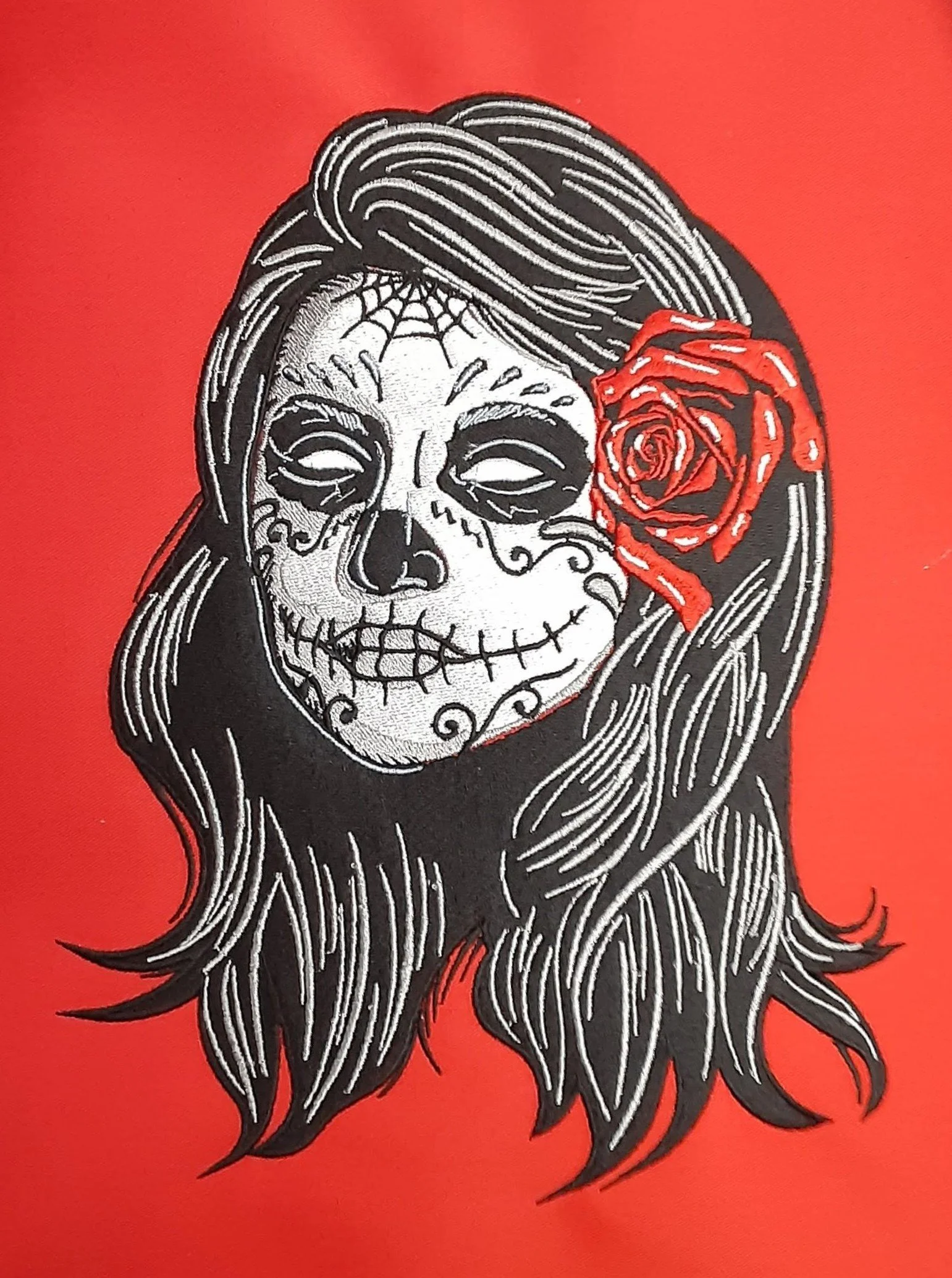 A detailed embroidery of a woman with a Day of the Dead sugar skull face paint, long hair, and a red rose in her hair, set against a red background.