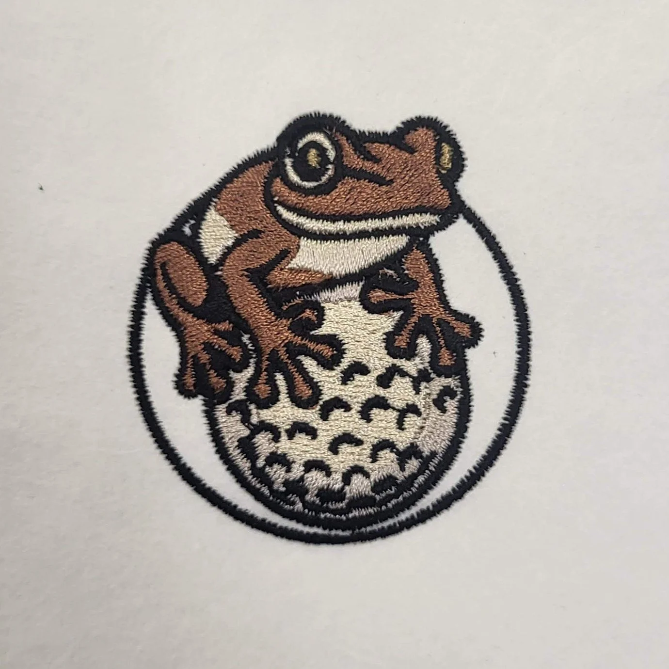 Embroidered image of a frog with brown and white coloring sitting on top of a golf ball, positioned on a white background.