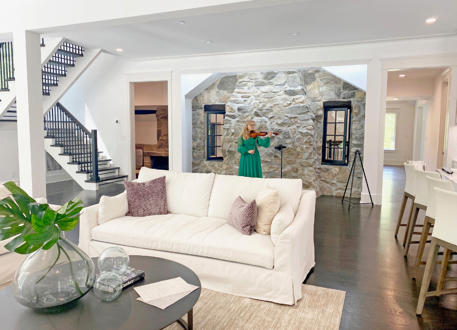 Modern living room with white sofa, decorative cushions, indoor plant in glass vase, and woman in green dress playing violin by stone wall.