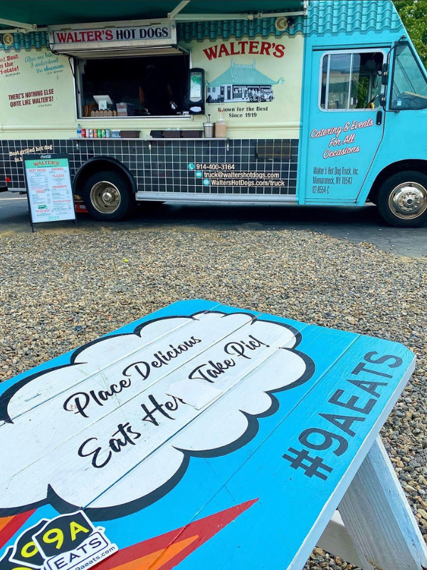 A vintage-style food truck named Walter's Hot Dogs with blue accents is parked on a gravel surface. It features signage for hot dogs and catering services. In the foreground, a painted picnic table displays the text 'Place Delicious Eats Here, Take Pic!' and the hashtag #9AEATS.
