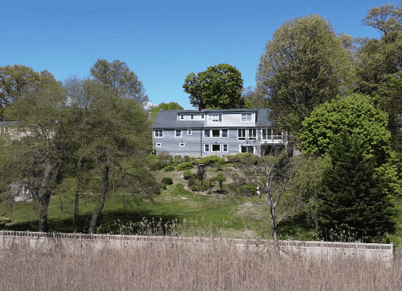 Large gray house surrounded by trees and greenery with a pond in the foreground, under a clear blue sky.