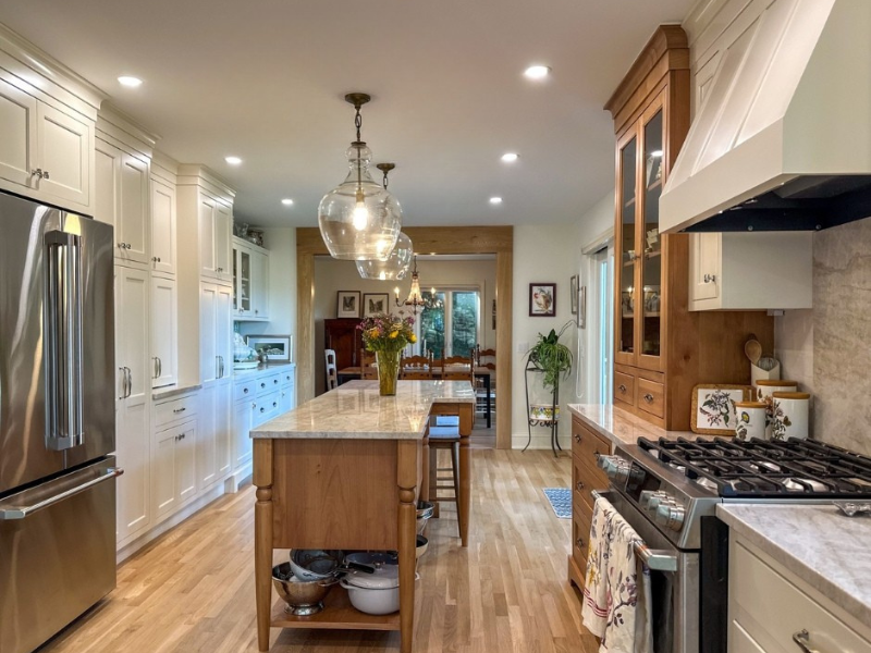 Kitchen with white cabinets, a large wooden island with a vase of flowers, pendant lights, stainless steel refrigerator, stove, and a dining area in the background with a table and chairs, hardwood floors, and trees visible through sliding glass door.