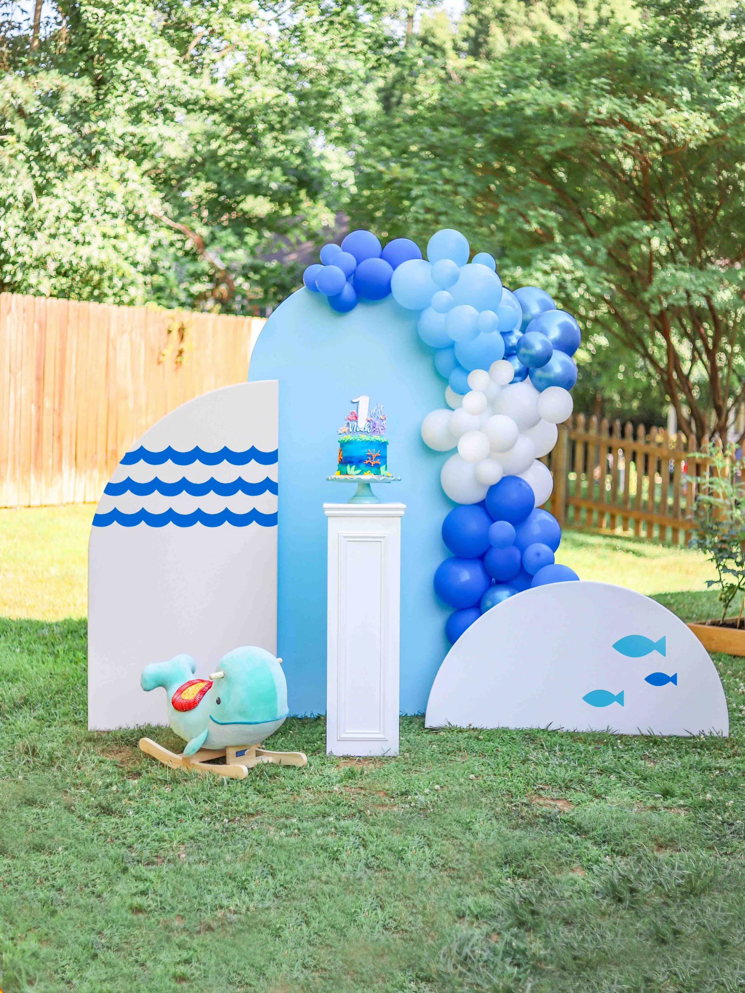 Underwater-themed birthday party decoration setup with a whale plush toy, blue and white balloons, and a cake on a pedestal with a '1' on top.