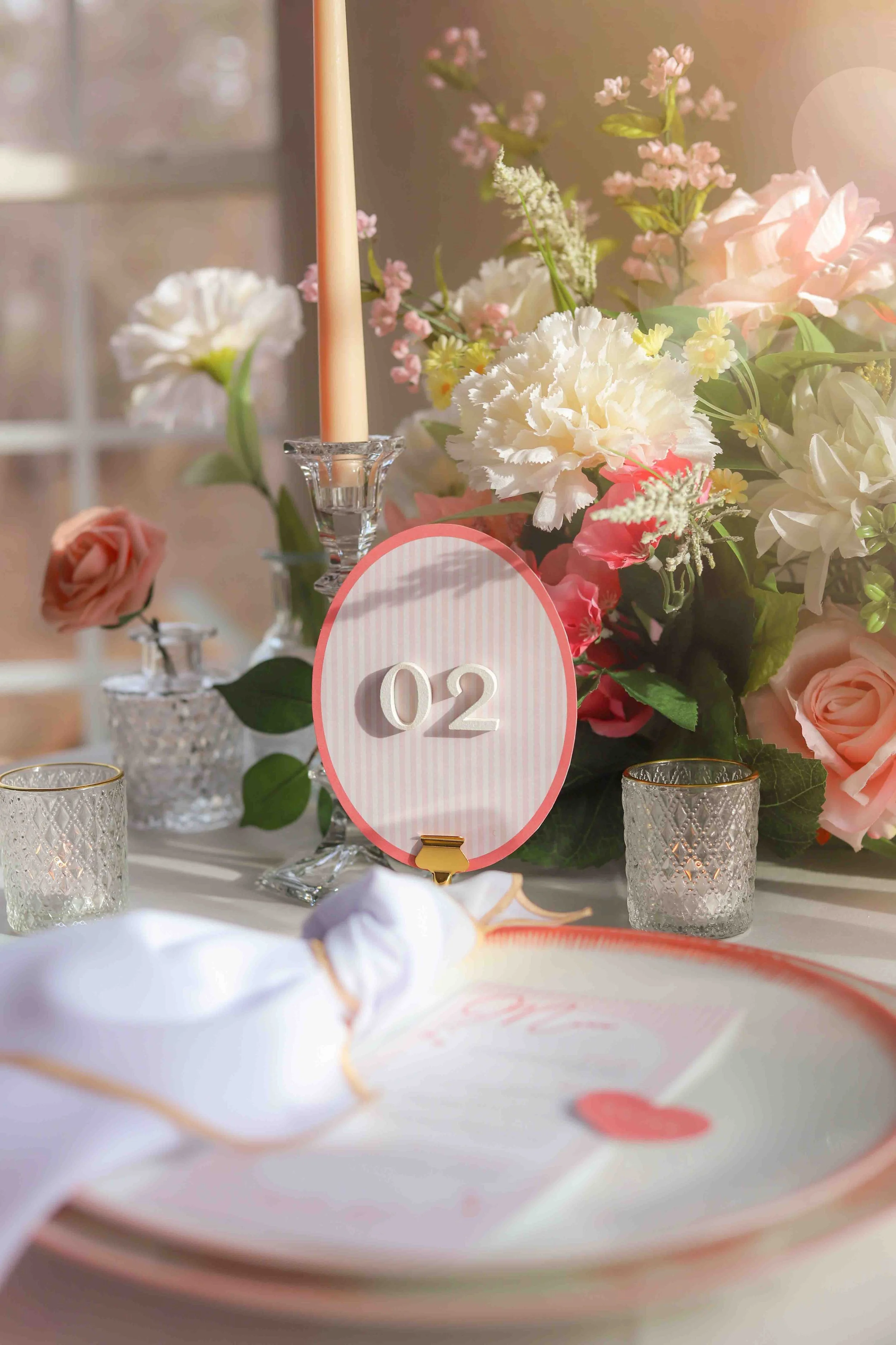 Table setting with a pink and white table number sign displaying '02', surrounded by candles and a floral arrangement of pink and white roses and other flowers.