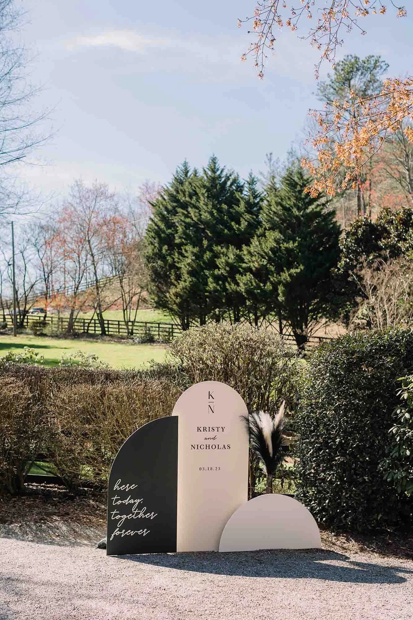 A decorative sign with the names Kristy and Nicholas, and the date 03.18.23, placed outdoors among bushes and trees under a clear sky.