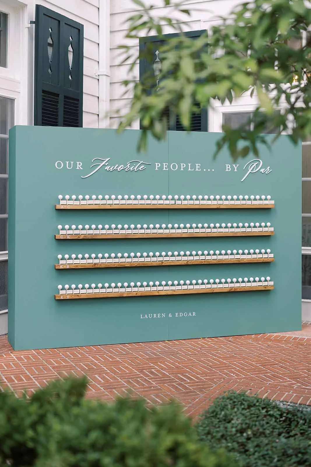 A wedding guestbook display with a teal backdrop and four horizontal wooden shelves holding small numbered white pegs, with a green shrub in the foreground and part of a house in the background. The sign reads 'Our Favorite People... by Dr' with the names 'Lauren & Edgar' at the bottom.