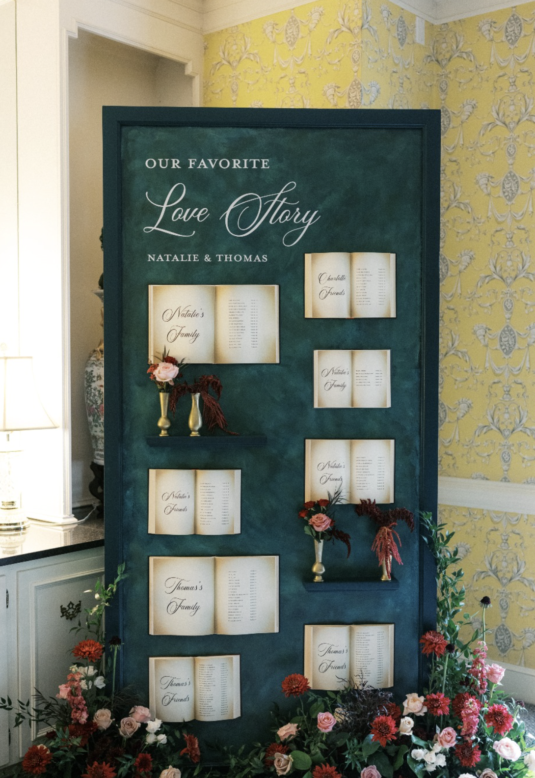 A display board with the title 'Our Favorite Love Story,' featuring open book pages with names and lists, decorated with vases of pink and red flowers, at a wedding reception or event.