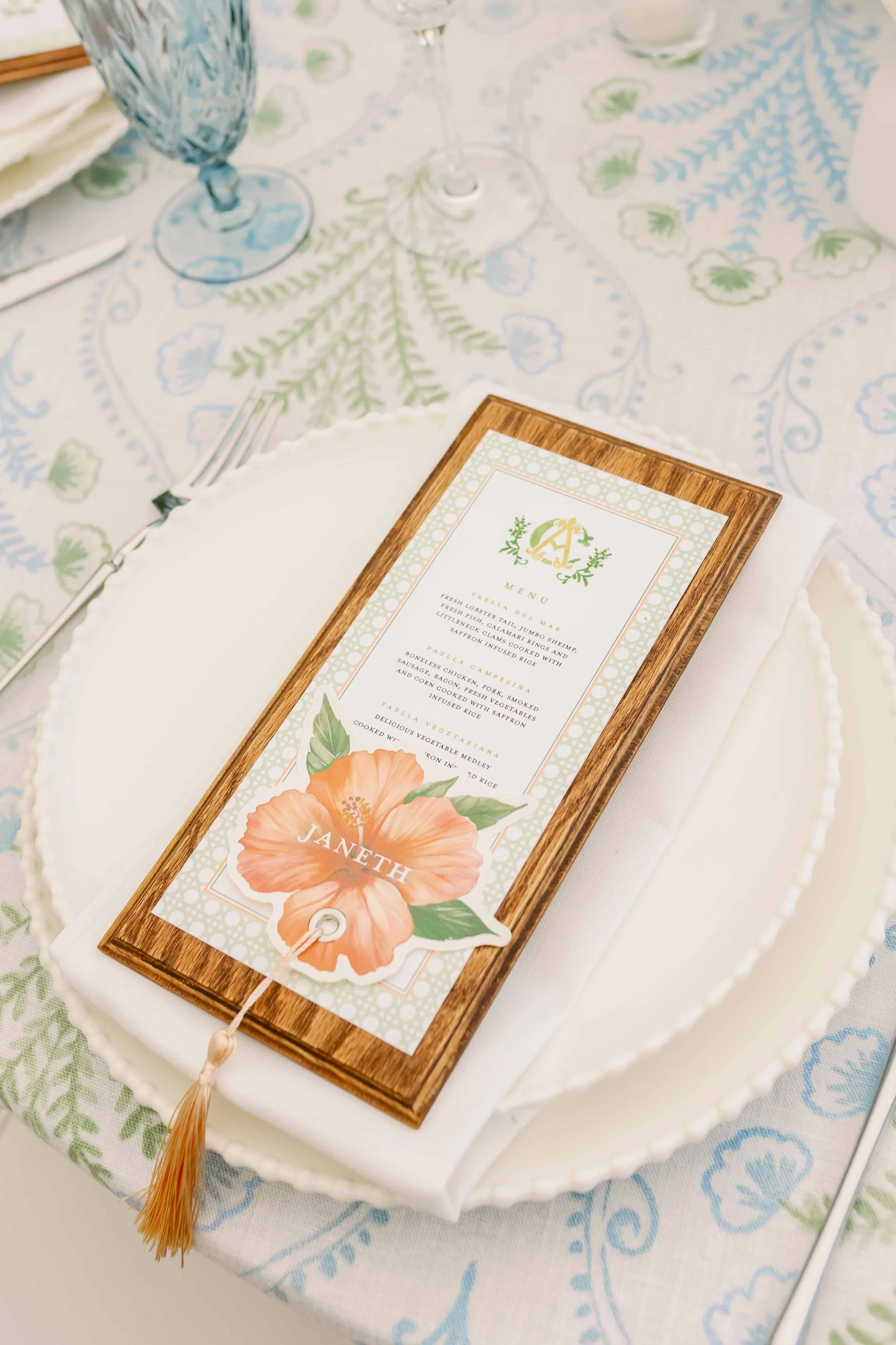 A place setting on a table with a white plate, a napkin, and a menu card. The menu card is framed in wood and decorated with a large orange hibiscus flower and green leaves, with the name "Janeth" on a smaller cutout. There are wine glasses and a tab