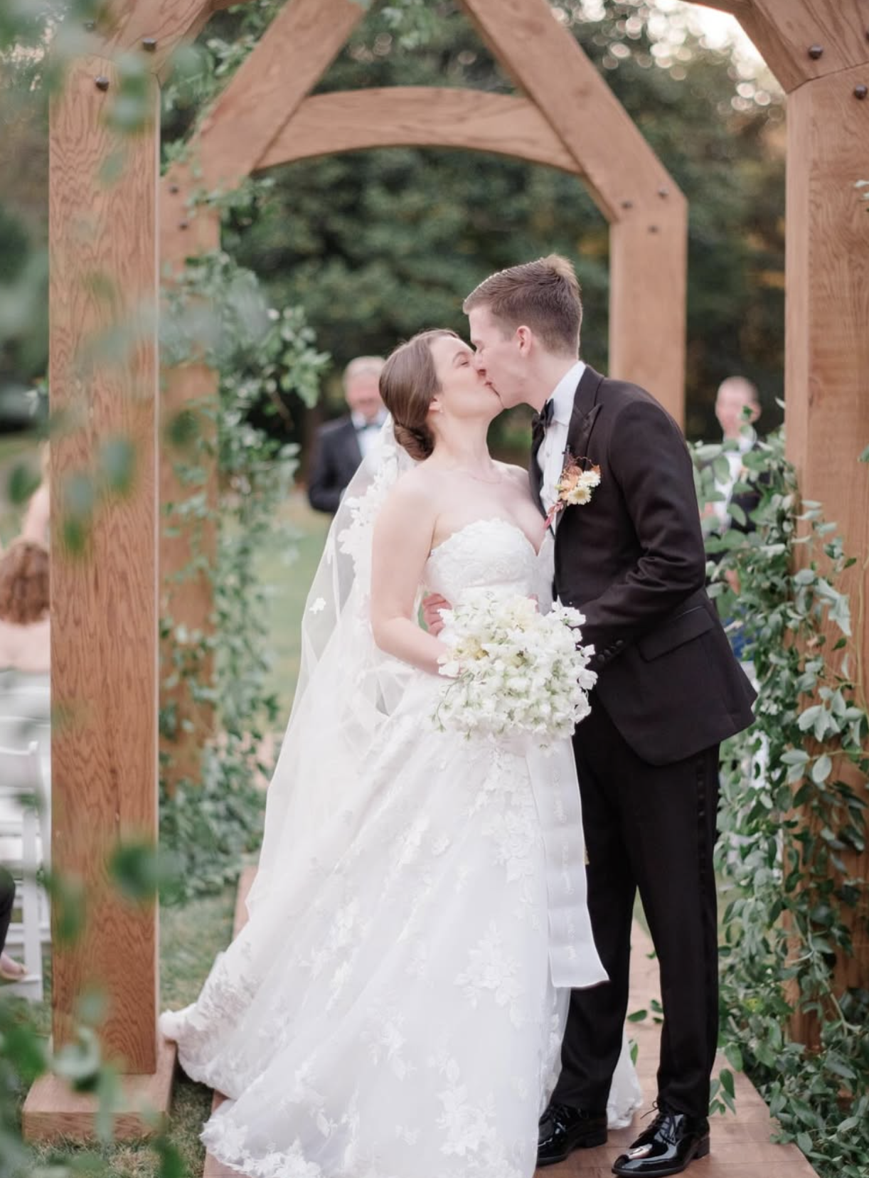 A bride and groom kiss during their outdoor wedding ceremony under a wooden arbor with green foliage.