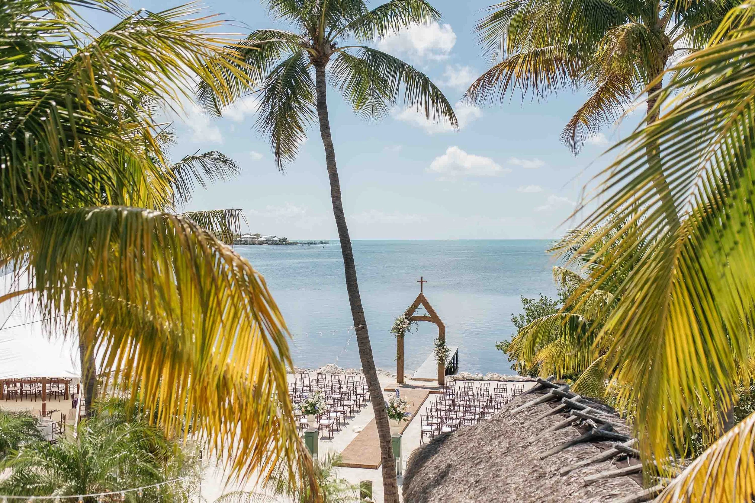 Beachside wedding setup with an arch decorated with flowers, chairs arranged facing the water, palm trees framing the scene, and the ocean in the background under a partly cloudy sky.
