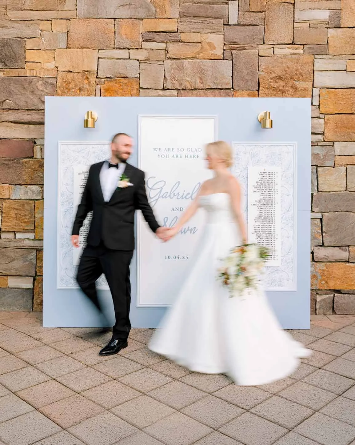 A bride and groom holding hands and smiling, standing in front of a wedding sign on a stone wall background.