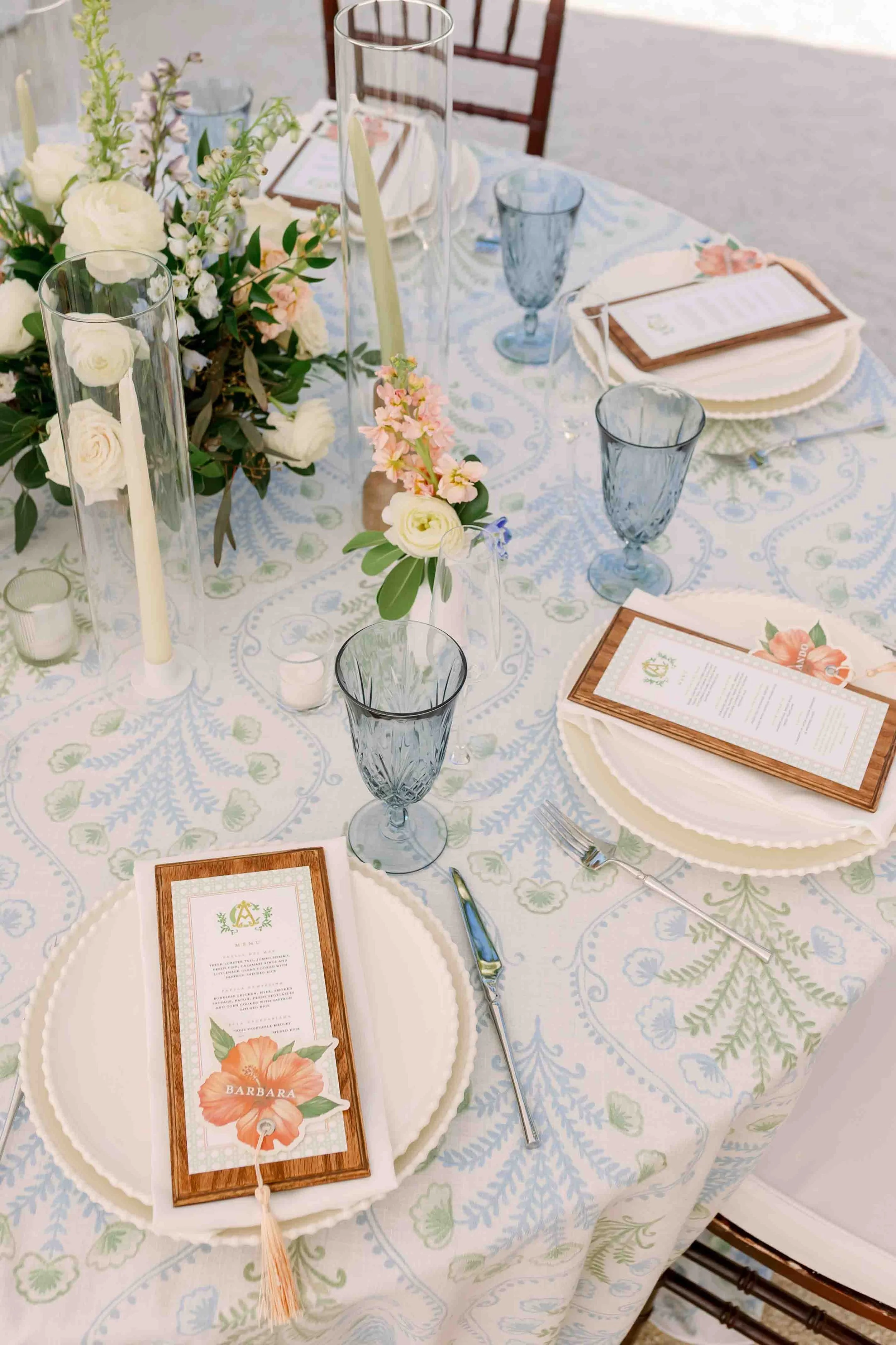 Elegant table setting with floral centerpiece, tall candles, menu cards, and blue glassware on a patterned tablecloth.