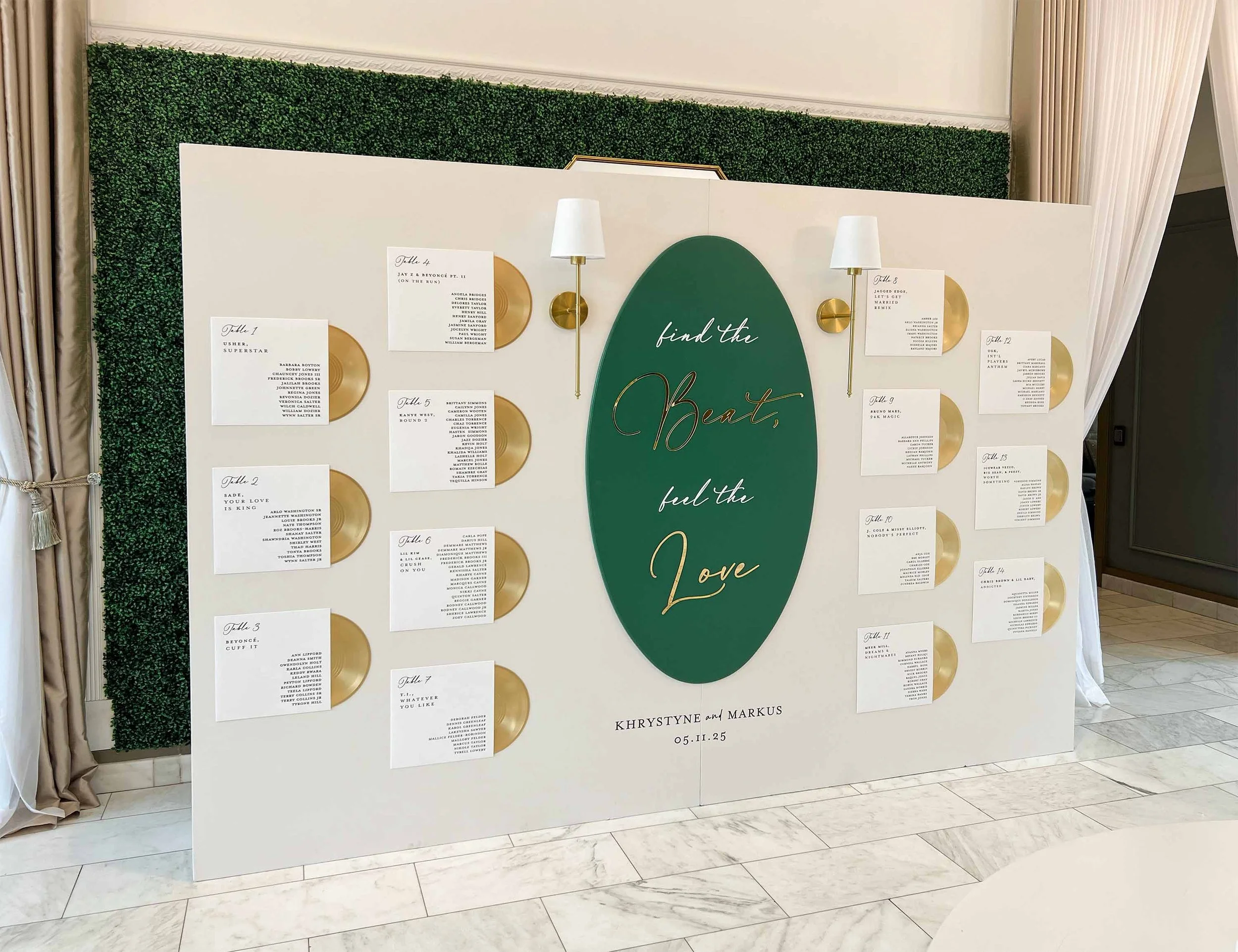 A seating chart display for a wedding, with 14 cards on a white board, each card listing guest names, adorned with gold plates and two wall lamps, with a green oval in the center containing the words "find the Beasts, feel the Love," and the names Kh