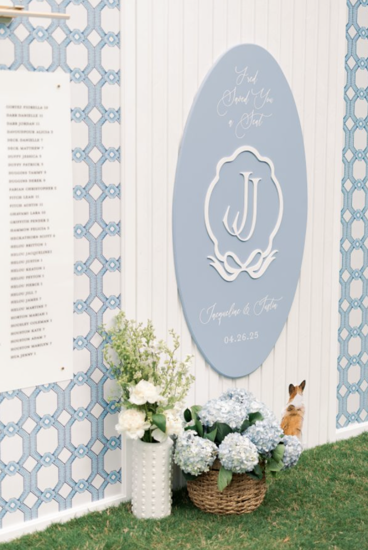 Decorative wedding display with a large blue oval sign featuring the monogram 'J' and the date '04.26.25', surrounded by white and blue flowers, a small pumpkin, and a dog with white and brown fur.