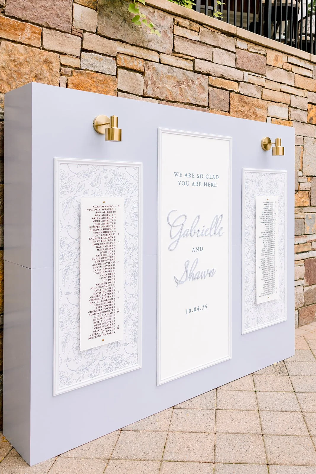 Wedding seating chart display with a white board, gold wall sconces, and floral border designs, showing the names Gabrielle and Shawn and the date 10.04.25.