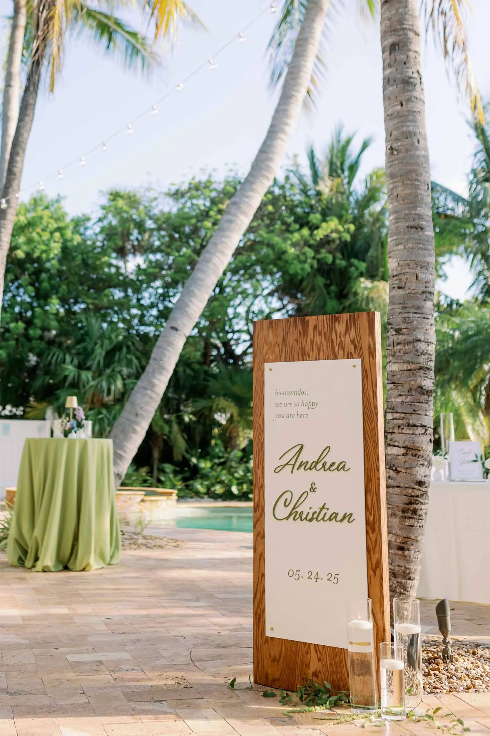 A wooden framed sign with elegant writing welcoming guests to Andrea and Christian's wedding on May 24-25, placed outdoors near palm trees and a pool, with tables decorated for the celebration.