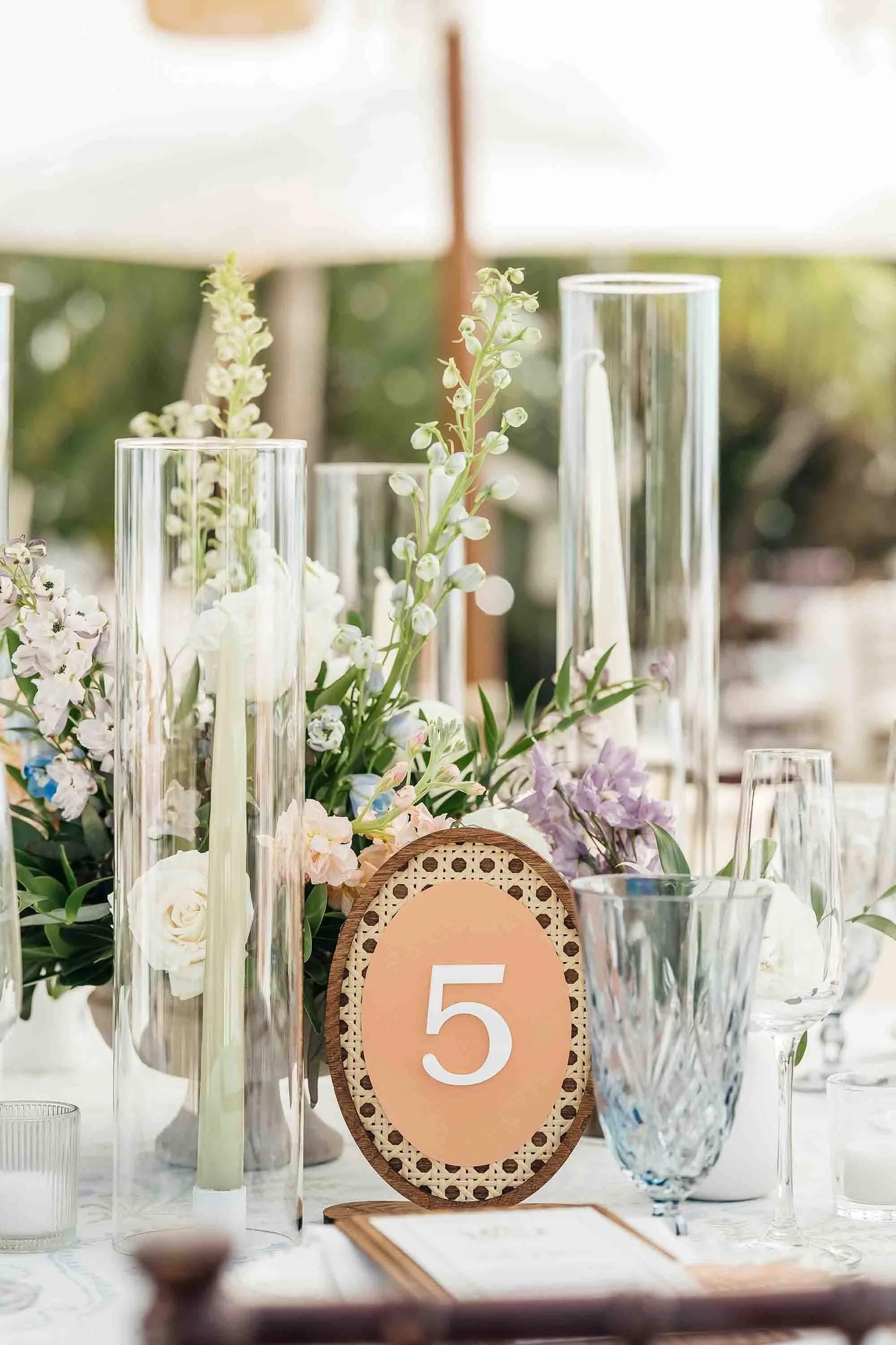 A table centerpiece with tall glass candle holders, a floral arrangement with white, purple, and blue flowers, a table number card with the number 5, and various drinking glasses, set outdoors under a white canopy.