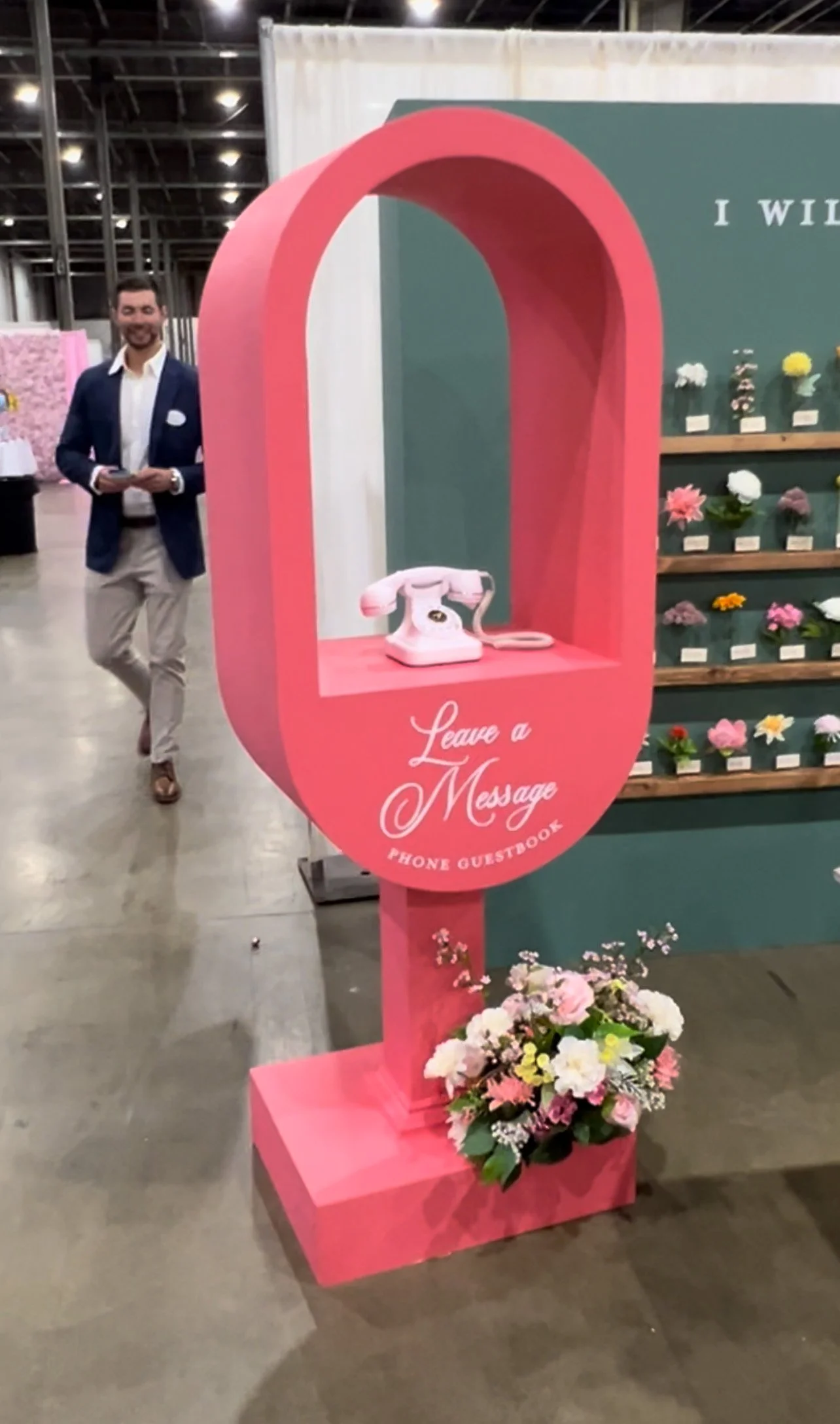 A pink phone booth with a vintage rotary phone inside, decorated with flowers at the base. The booth has the text 'Leave a Message' and 'Phone Guestbook' on it. In the background, a man in a blue blazer and beige pants is walking past in a large indo