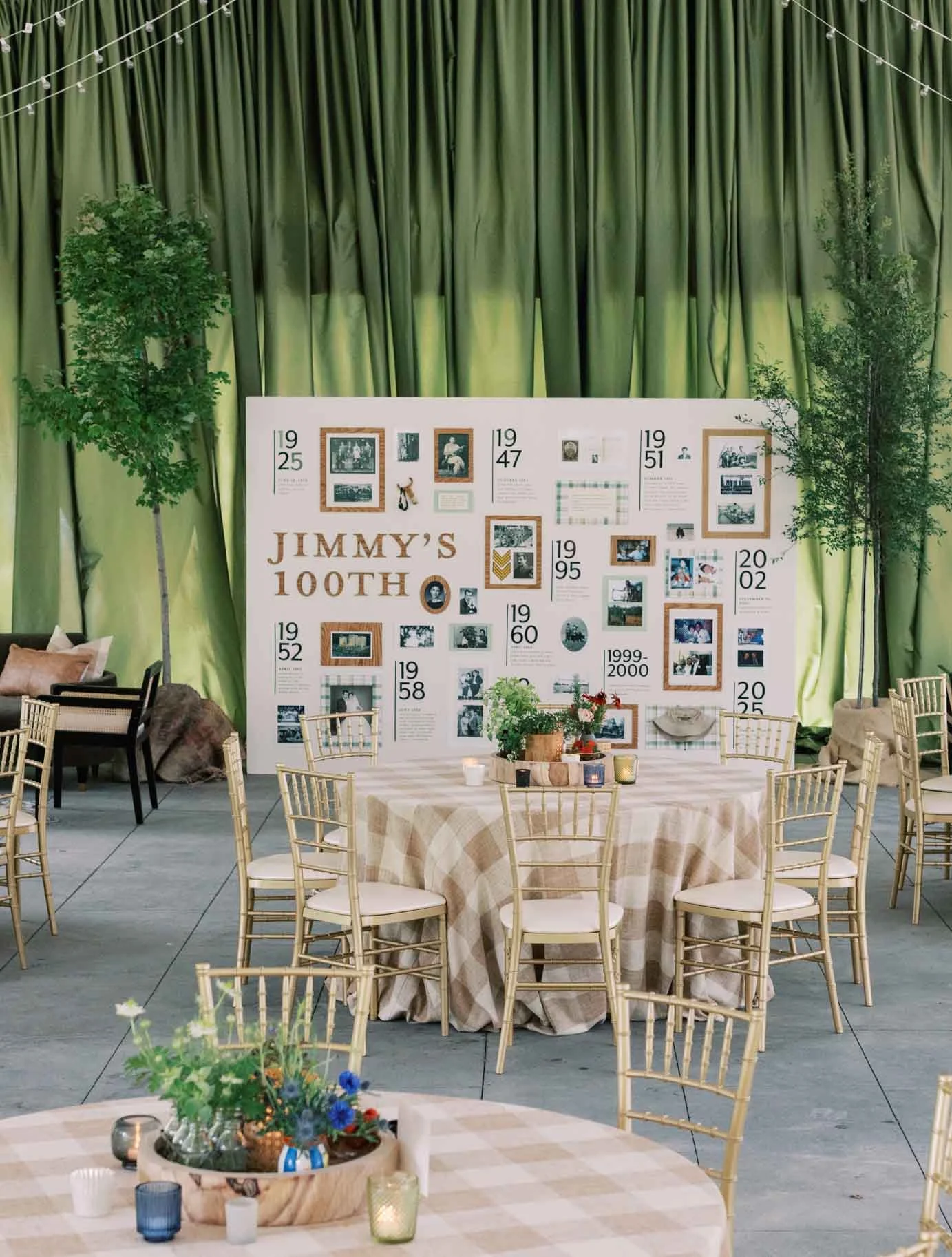 Decorative display with a large white board commemorating Jimmy's 100th birthday, surrounded by tables with checkered tablecloths, chairs, candles, and floral arrangements. Green curtains and potted trees decorate the background.