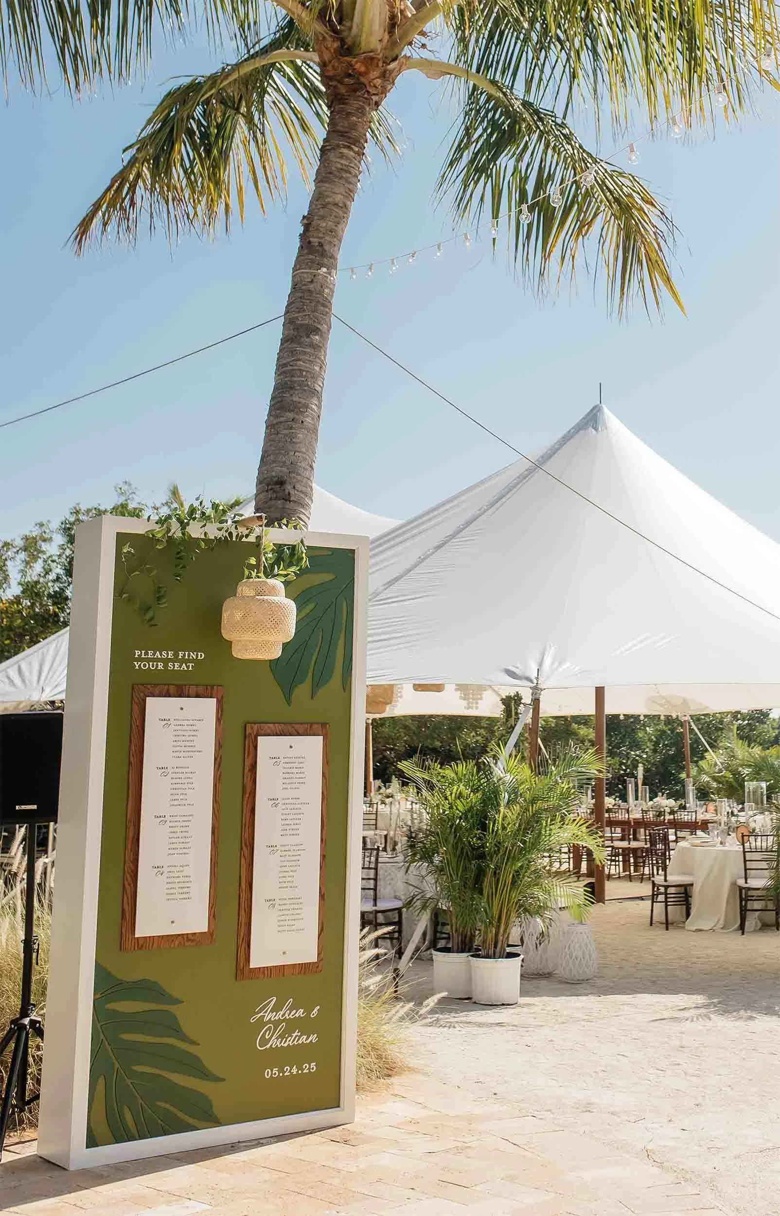 An outdoor wedding reception setup with a large white tent, tropical plants, a palm tree, and a green seating chart display with wooden frames, hanging lantern, and the names 'Andrea & Christian' along with the date '05.24.25'.