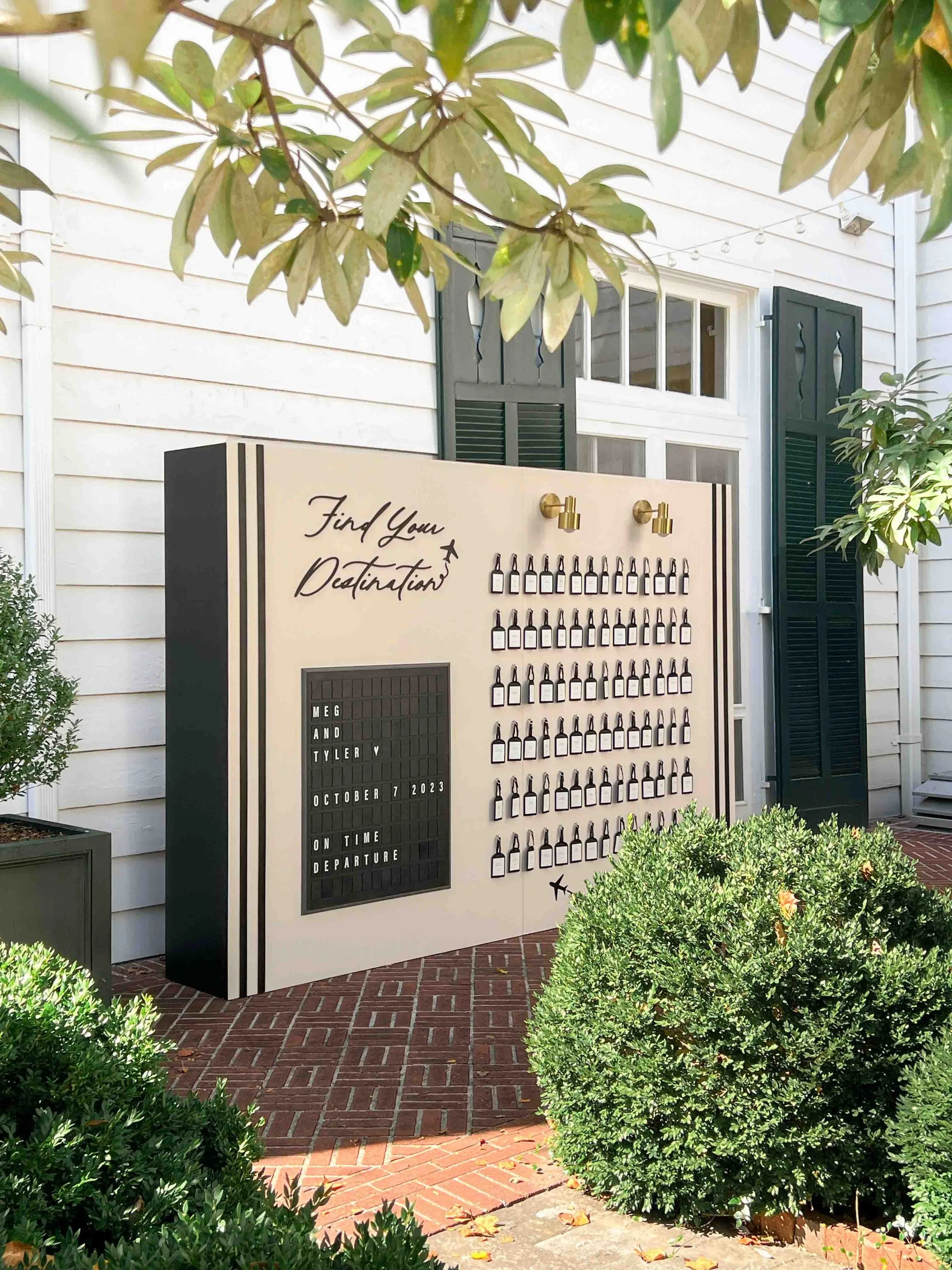 A decorative display board outdoors with the message "Find Your Destination" in cursive, a list of small bottles, and a message stating "Meg and Tyler, October 7, 2023, on time departure." The display is surrounded by greenery including bushes and tr