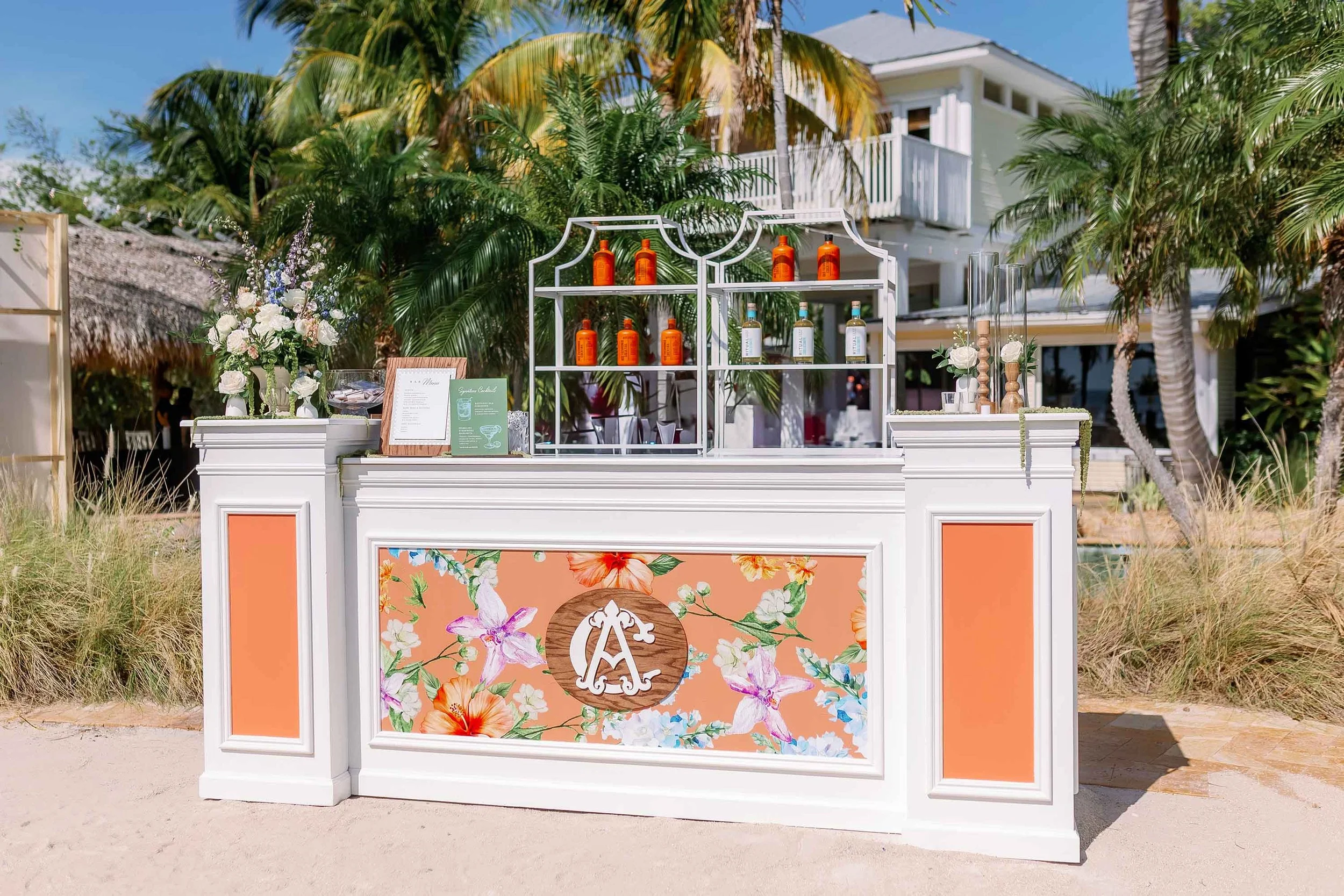 A beachside bar with white furniture and floral accents, set against palm trees and a house, featuring bottles of colorful liquor and a floral sign with initials.