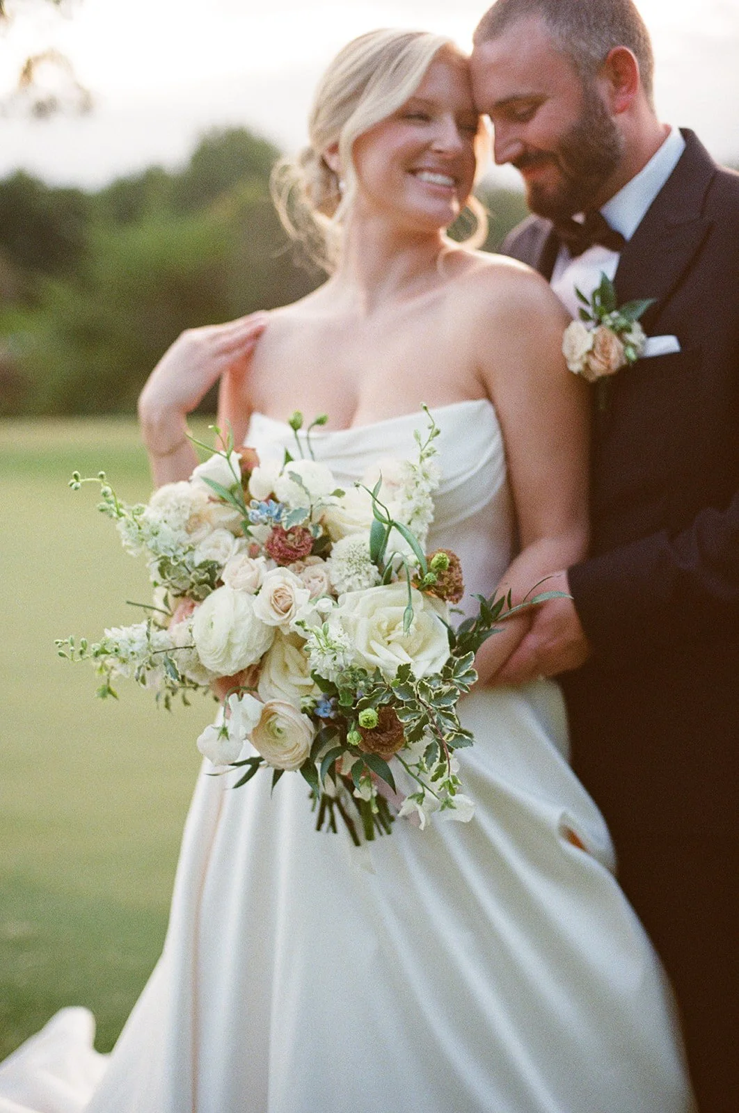 A bride and groom standing close, smiling and touching foreheads outdoors, with the bride holding a large bouquet of white and pastel flowers, on a grassy field during sunset.