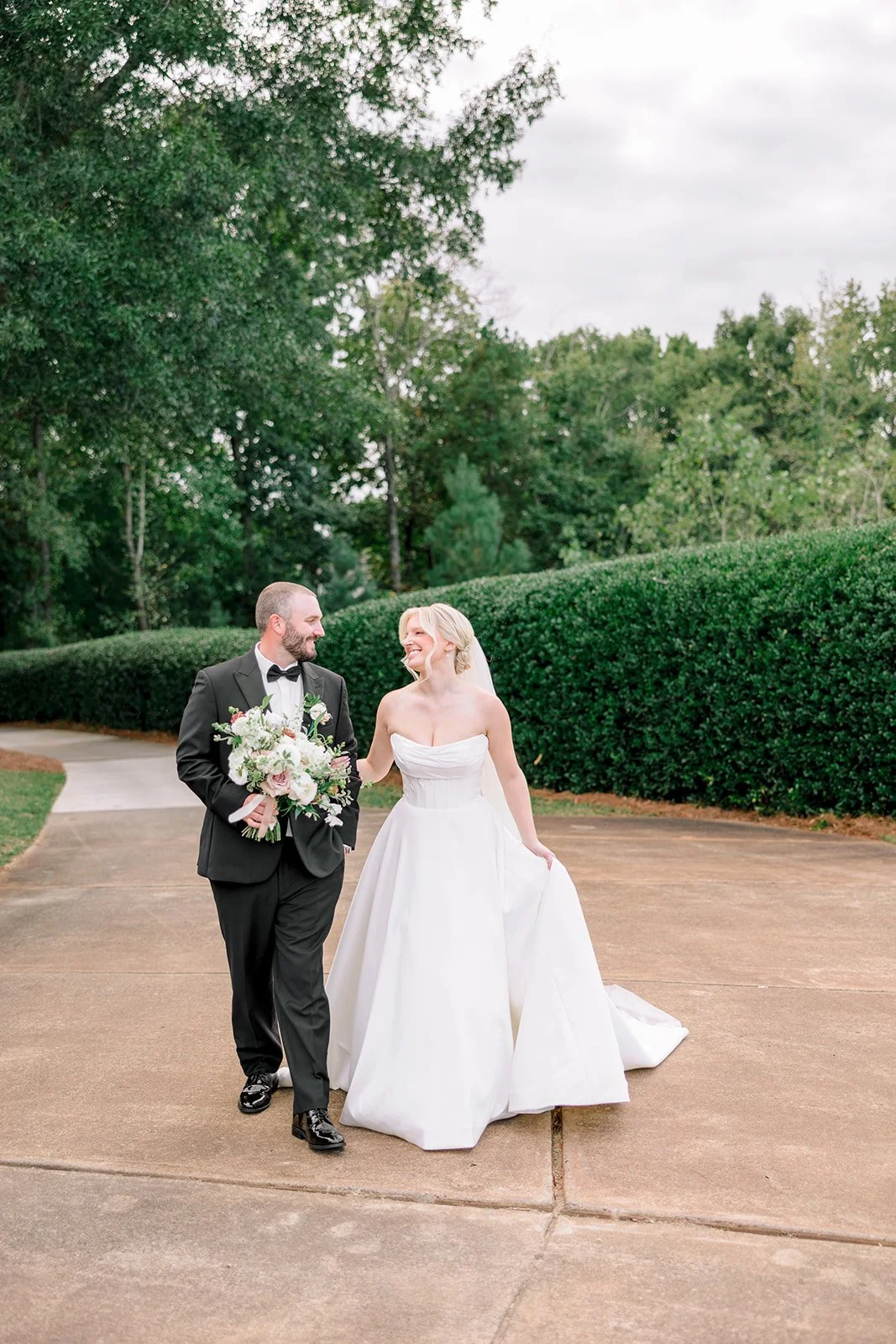 A bride in a white wedding gown and a groom wearing a black tuxedo walk together outdoors, smiling at each other, with a landscaped background of green trees and bushes.