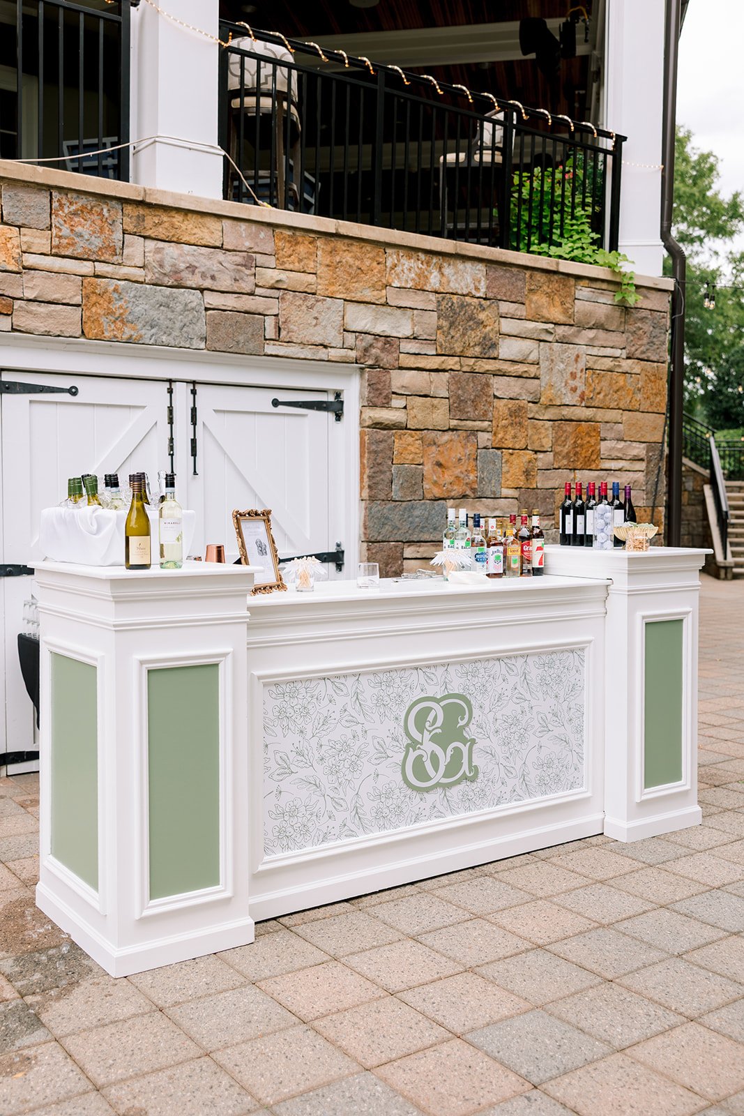 Outdoor bar setup with bottles of alcohol, wine, and mixers on a white counter against a brick wall, with string lights overhead.
