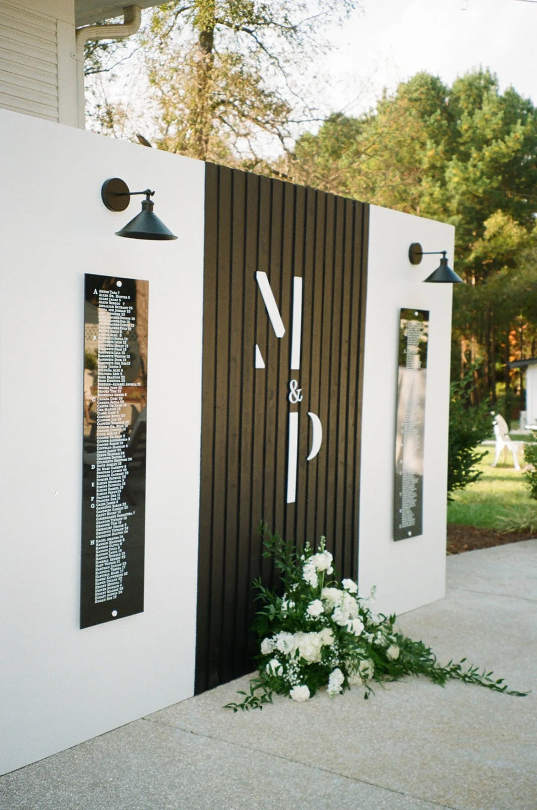 Exterior wall with Nashville-themed decor, including black and white vertical signs with text, black wall-mounted lamps, and a flower arrangement at the base, set in a lush, green outdoor environment.
