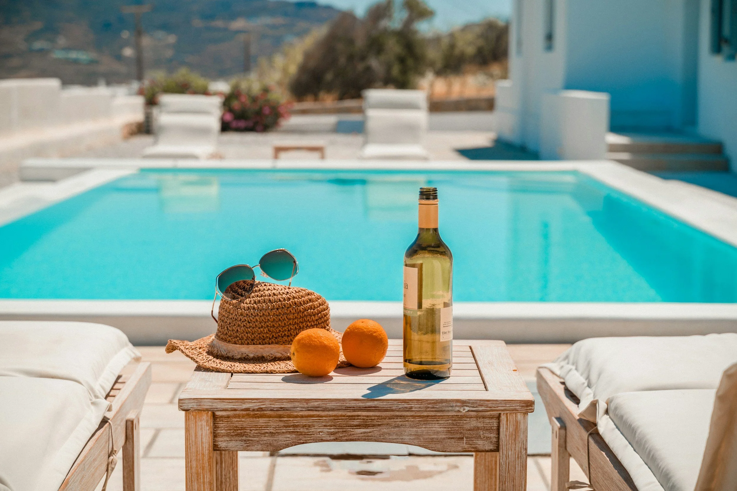 A lounge chair with a table next to a swimming pool, with a bottle of white wine, a straw hat, sunglasses, and two oranges on the table, in a sunny outdoor setting.
