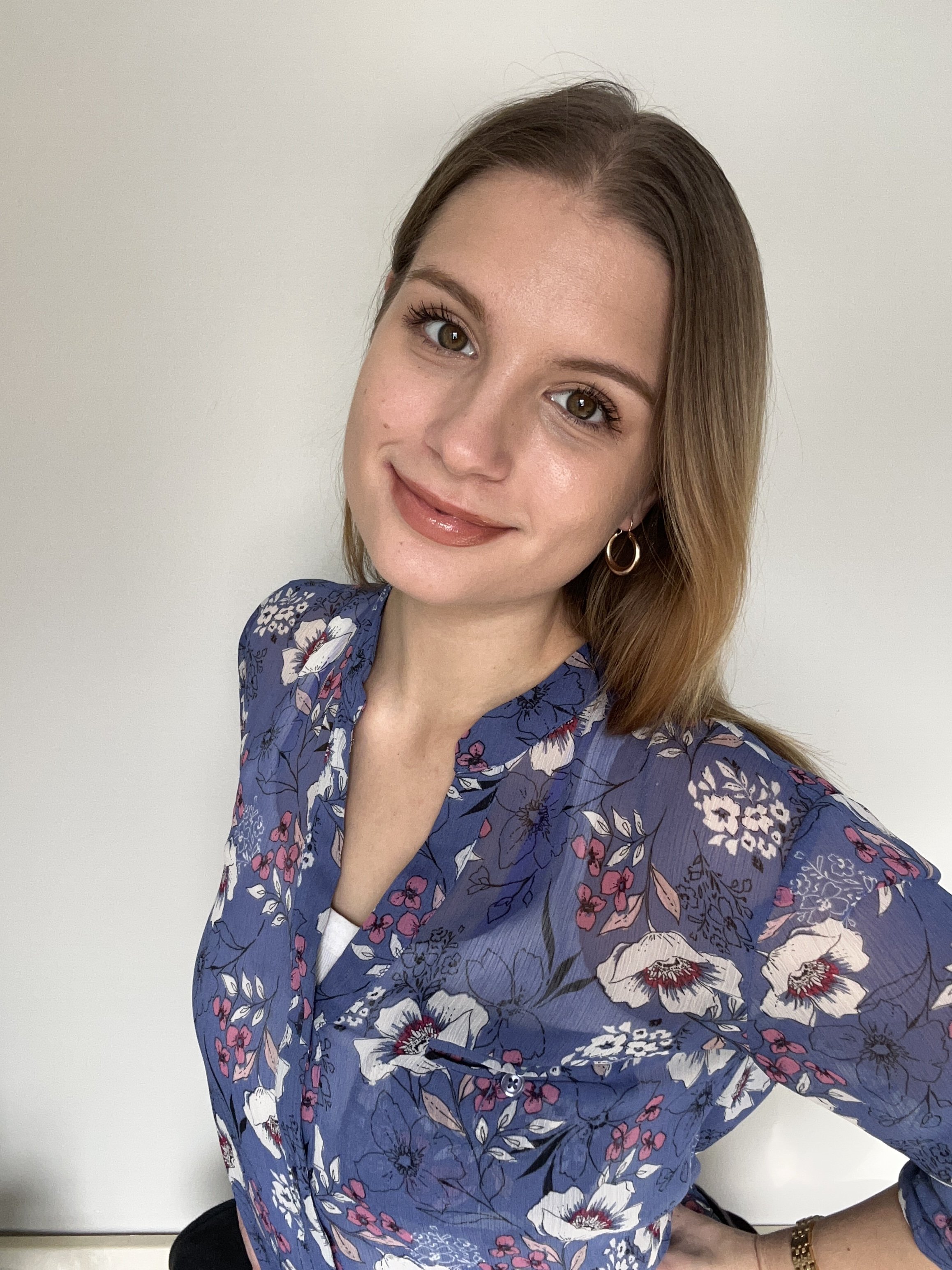A woman with light brown hair and brown eyes smiling at the camera, wearing a blue floral blouse and hoop earrings, standing against a plain light-colored wall.