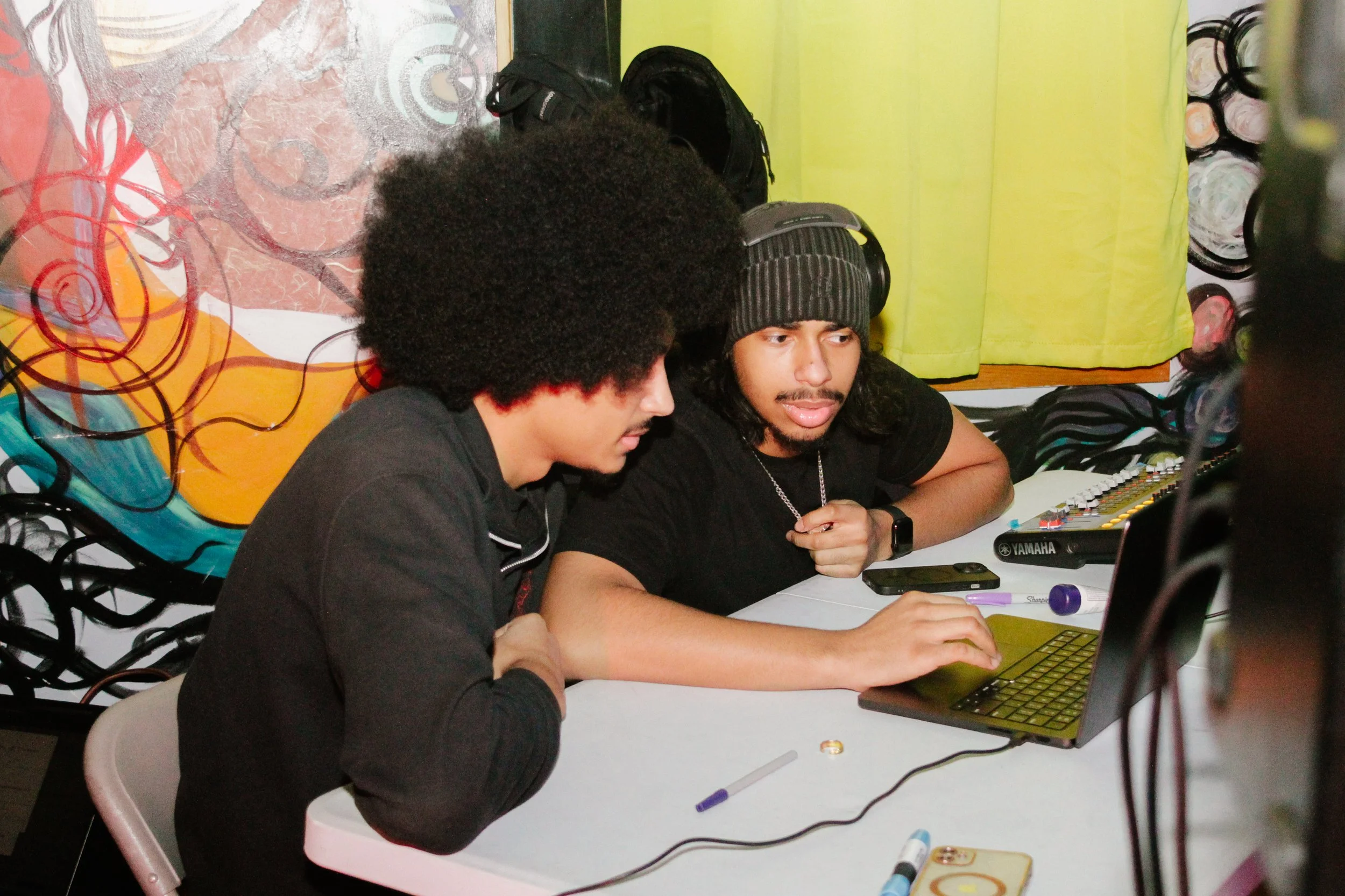 Two young men sitting at a table, working on a laptop, with art and yellow curtains in the background.